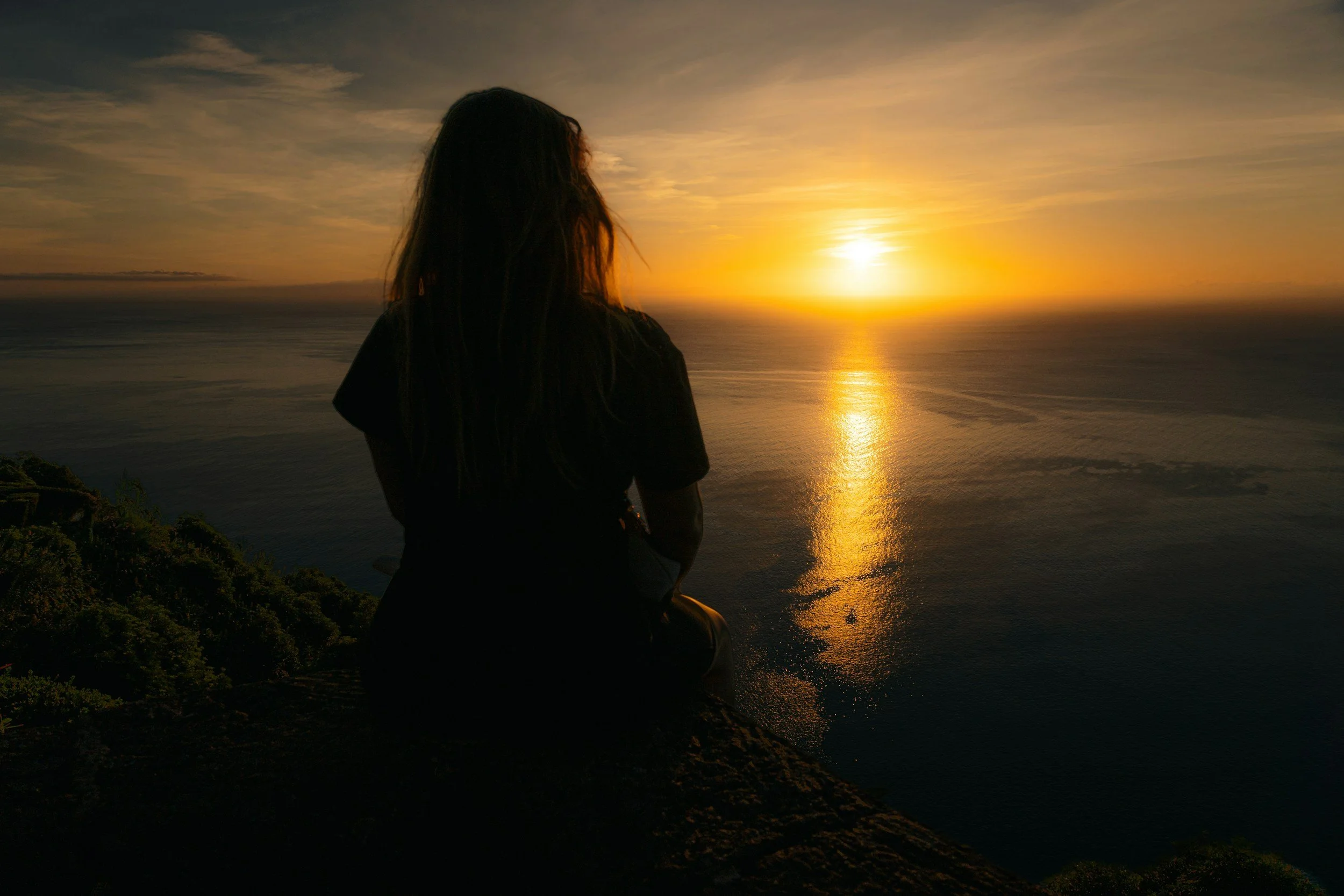 A person sitting on a hilltop watching the sunset over the ocean.