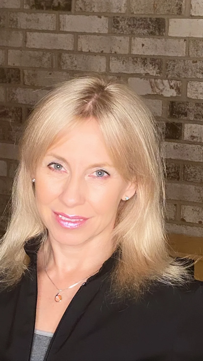 A smiling blonde woman with blue eyes wearing a black top or jacket, earrings, and a necklace, standing in front of a brick wall.