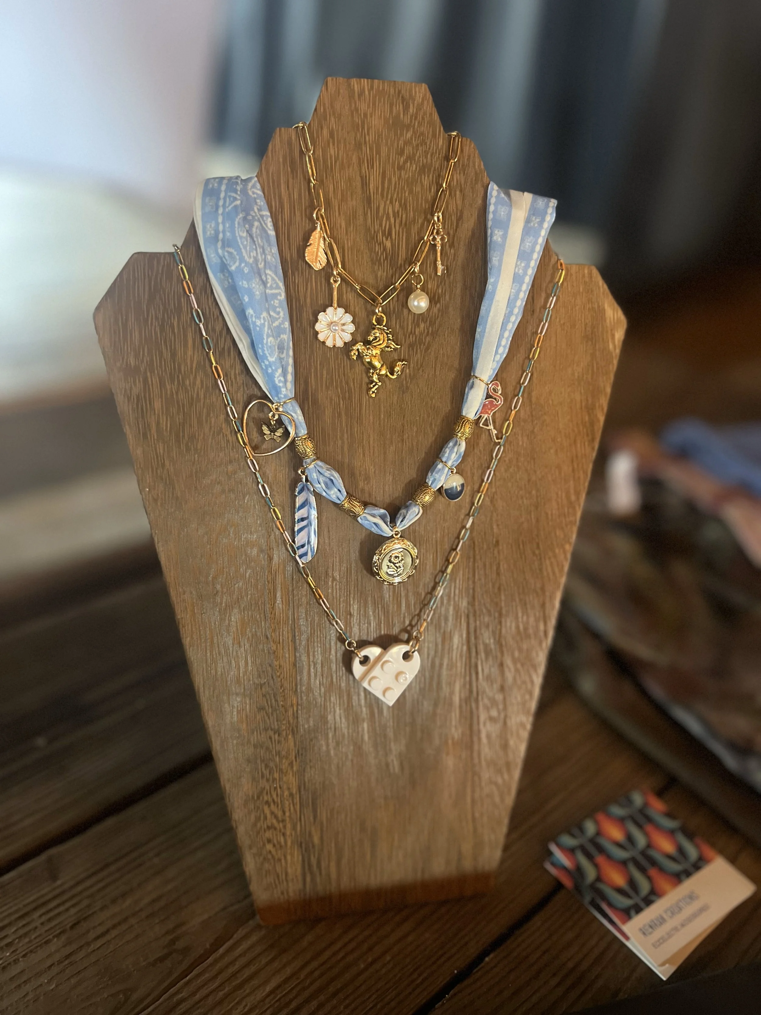 Layered necklaces with charms, including a daisy, a unicorn, a heart with a hoop, a flamingo, and a LEGO heart, displayed on a wooden jewelry stand.