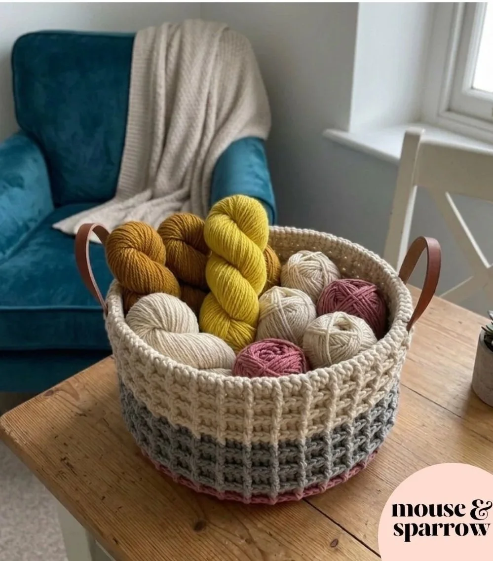 Crochet waffle stitch basket full of yarn on a white wooden table by a windwo