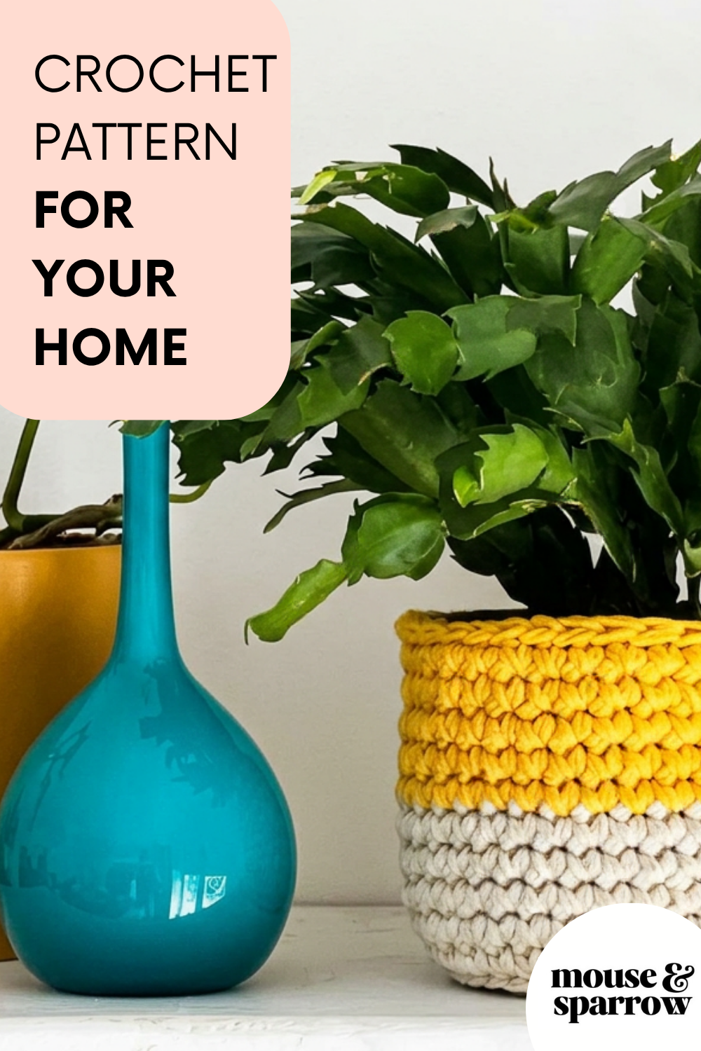 Crochet plant pot cover in golden yellow and natural cotton yarn, holding a Christmas cactus on a white shelf beside a teal glass vase — handmade home decor from Mouse and Sparrow.
