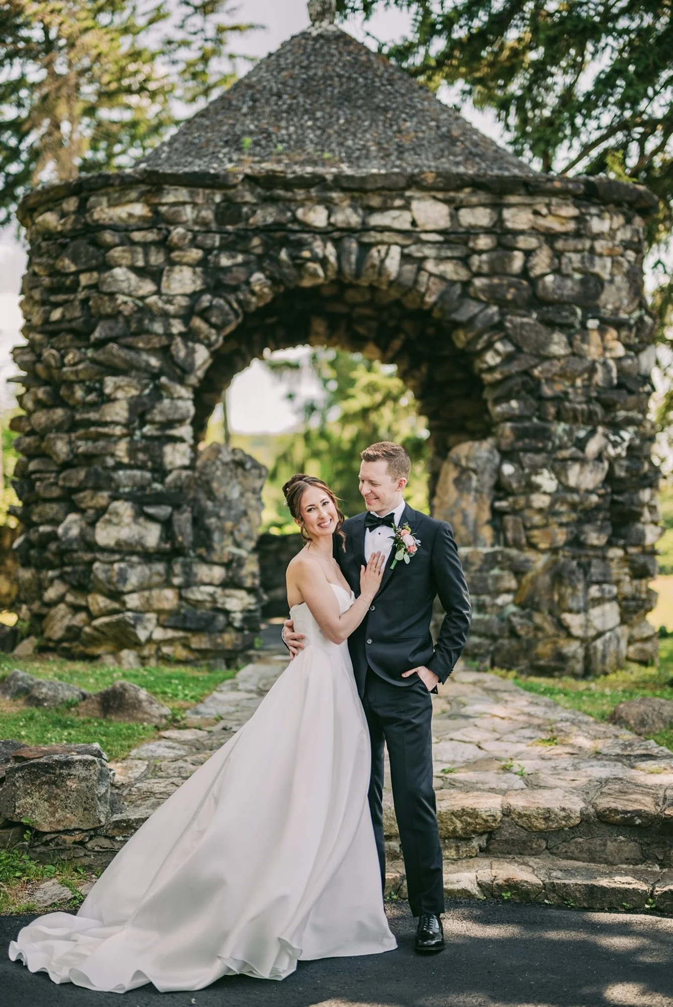 A bride and groom standing together outdoors in front of a stone archway, with the bride wearing a strapless white wedding gown with a long train and the groom in a black suit with a boutonniere, smiling and looking at each other.