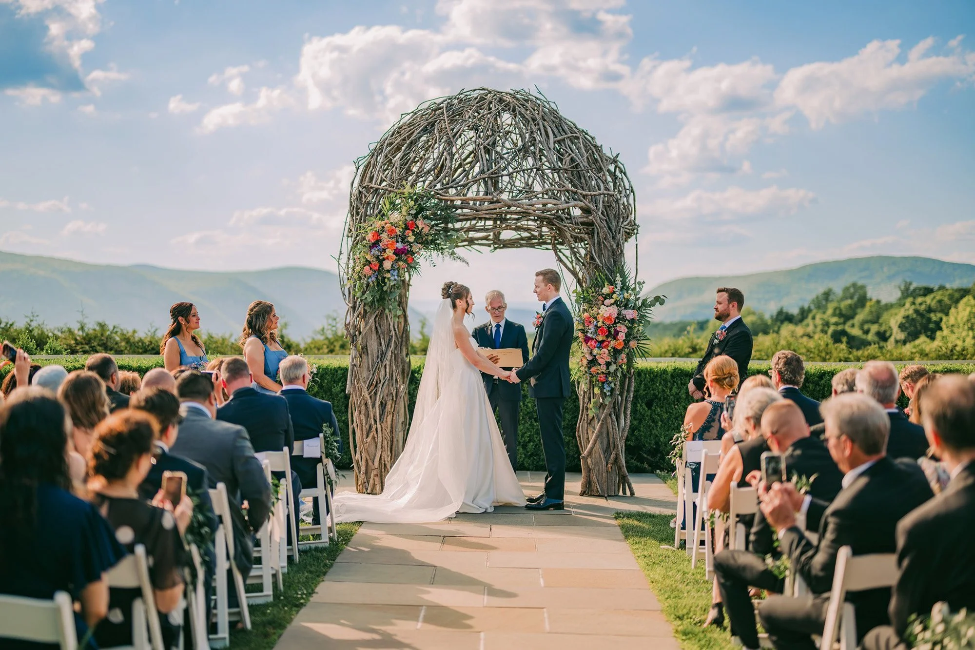 Outdoor wedding ceremony with bride and groom exchanging vows under a rustic wooden arch decorated with flowers, surrounded by seated guests with scenic mountains and sky in the background.