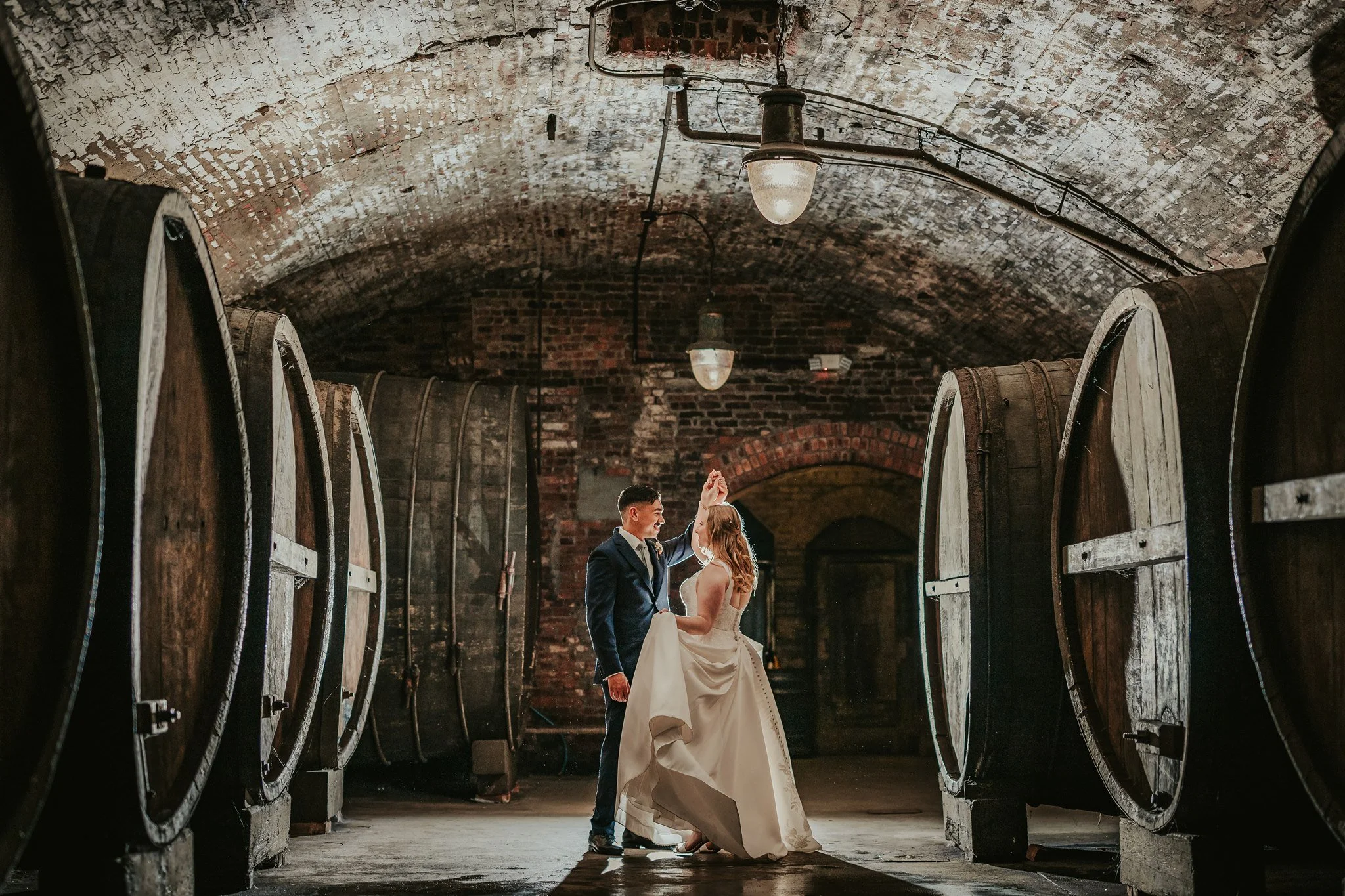 A bride and groom dancing in a rustic wine cellar with large wooden wine barrels on either side and exposed brick walls with arched brick ceiling.
