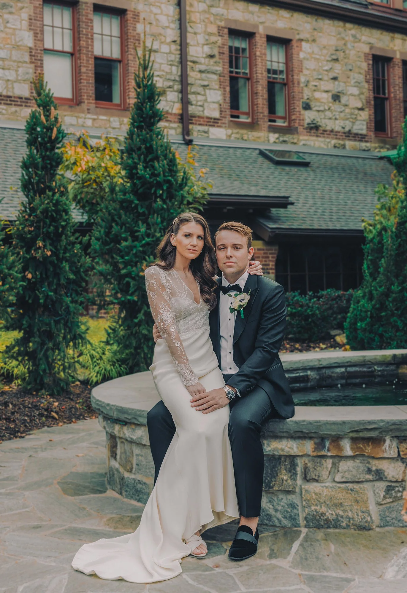 A bride and groom sitting together outdoors in front of a stone building with large windows, surrounded by greenery. The bride is wearing a long white wedding dress with lace details, and the groom is in a black tuxedo with a bow tie.