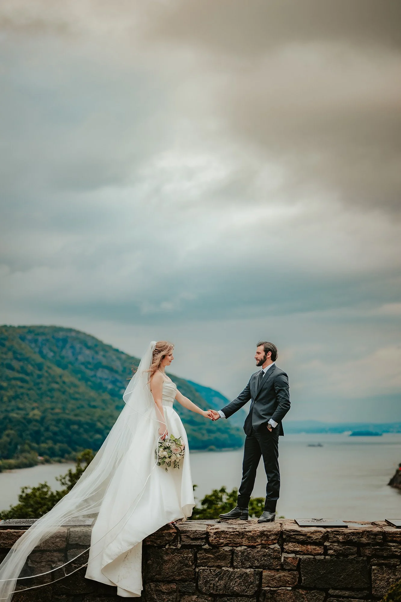 A bride and groom holding hands outdoors, standing on a stone wall with a lake and forested hills in the background. The bride is in a white wedding gown with a veil, holding a bouquet, while the groom is in a dark suit. The sky is overcast.