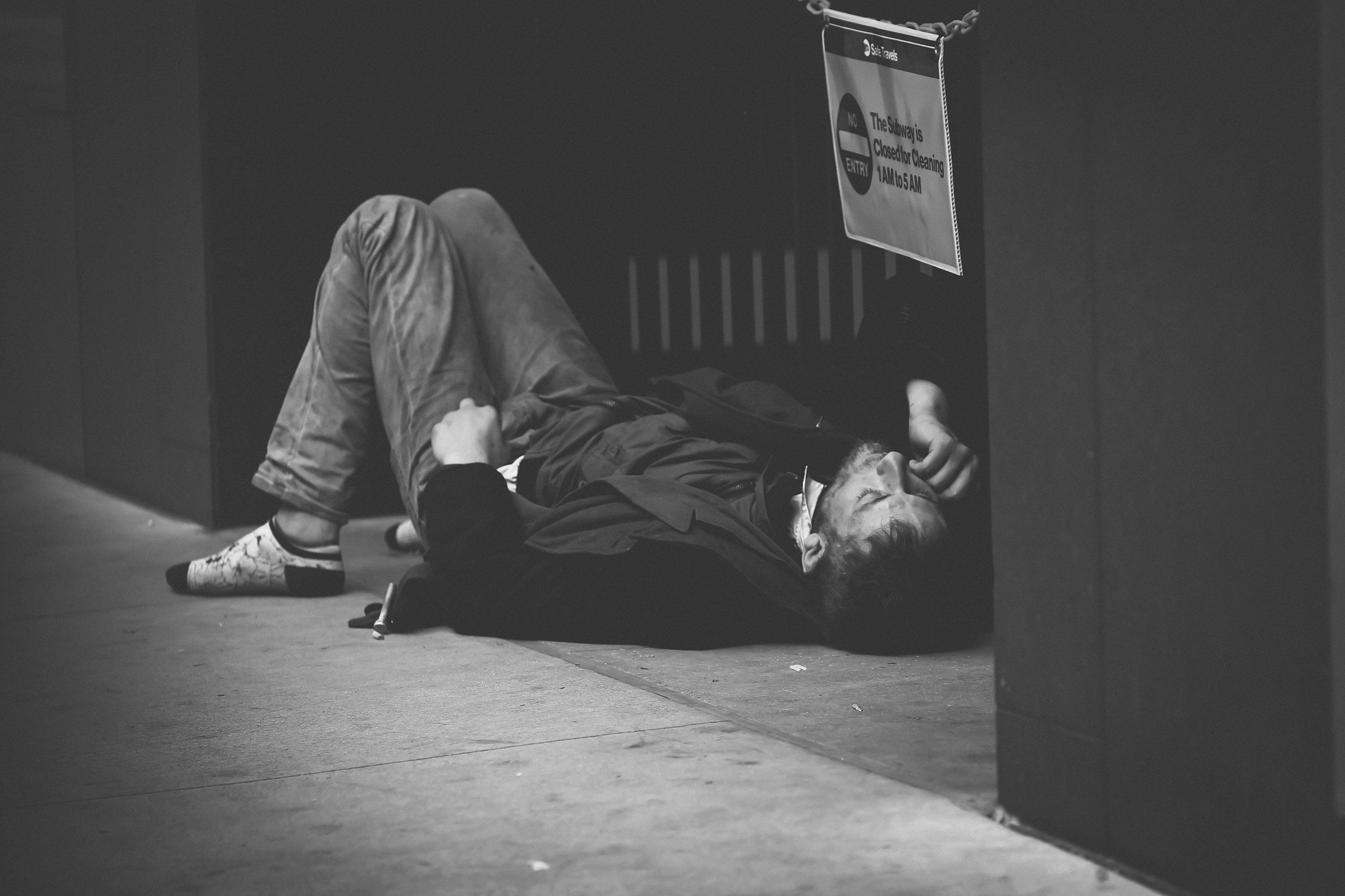 A person lying on the floor in a subway station, appearing to be asleep or unconscious, with a sign above indicating subway closure and cleaning hours.