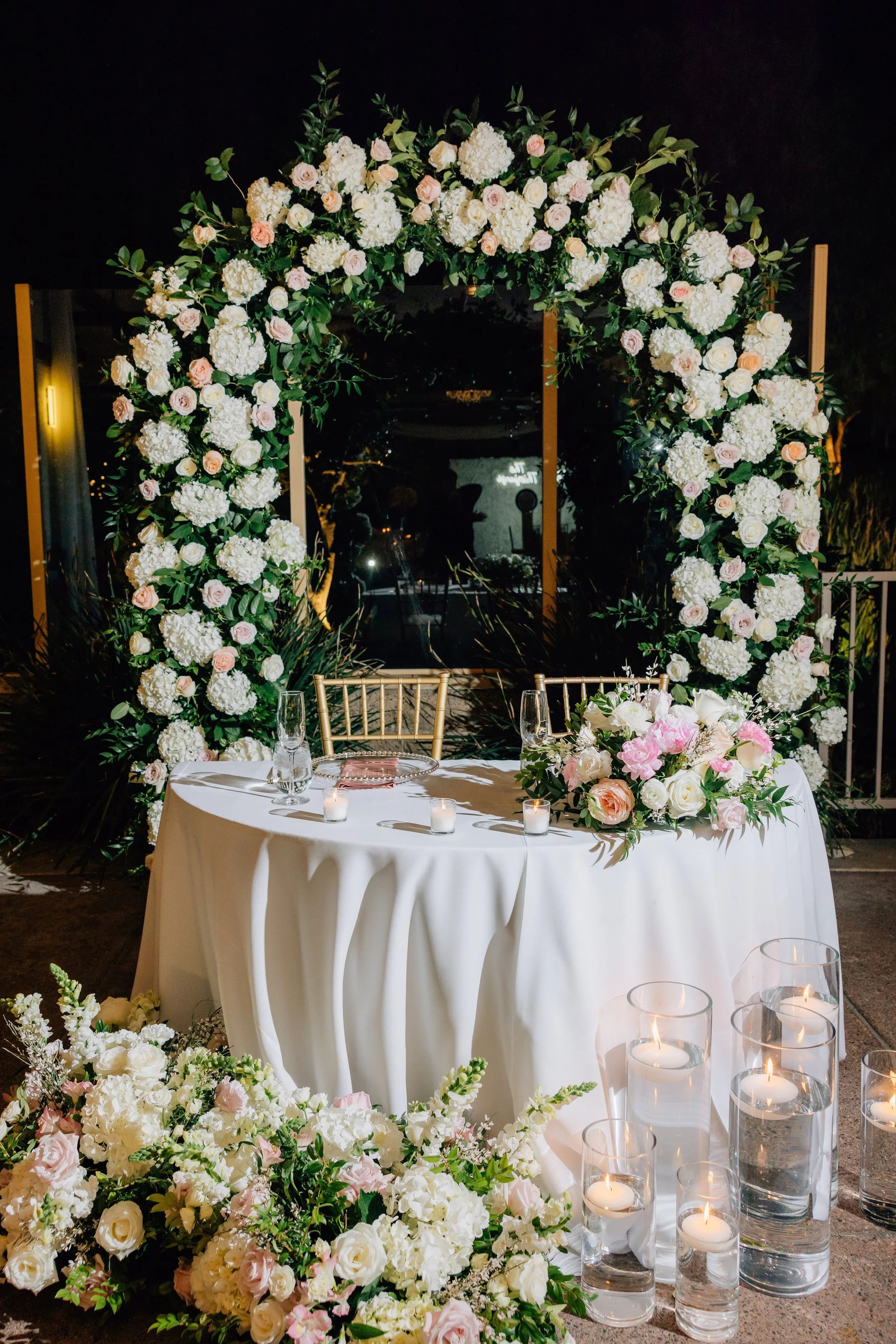 A happy couple at their wedding reception, standing behind a table under a floral arch, holding a glass of champagne.
