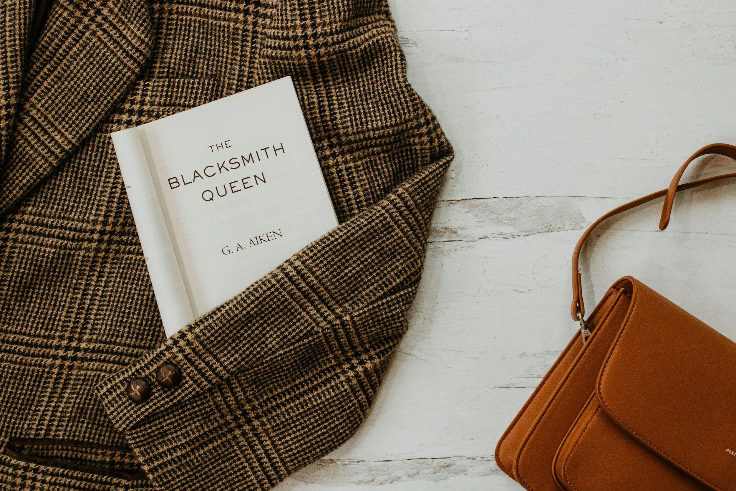 Title of book displayed with a plaid jacket and a tan purse on a marble background