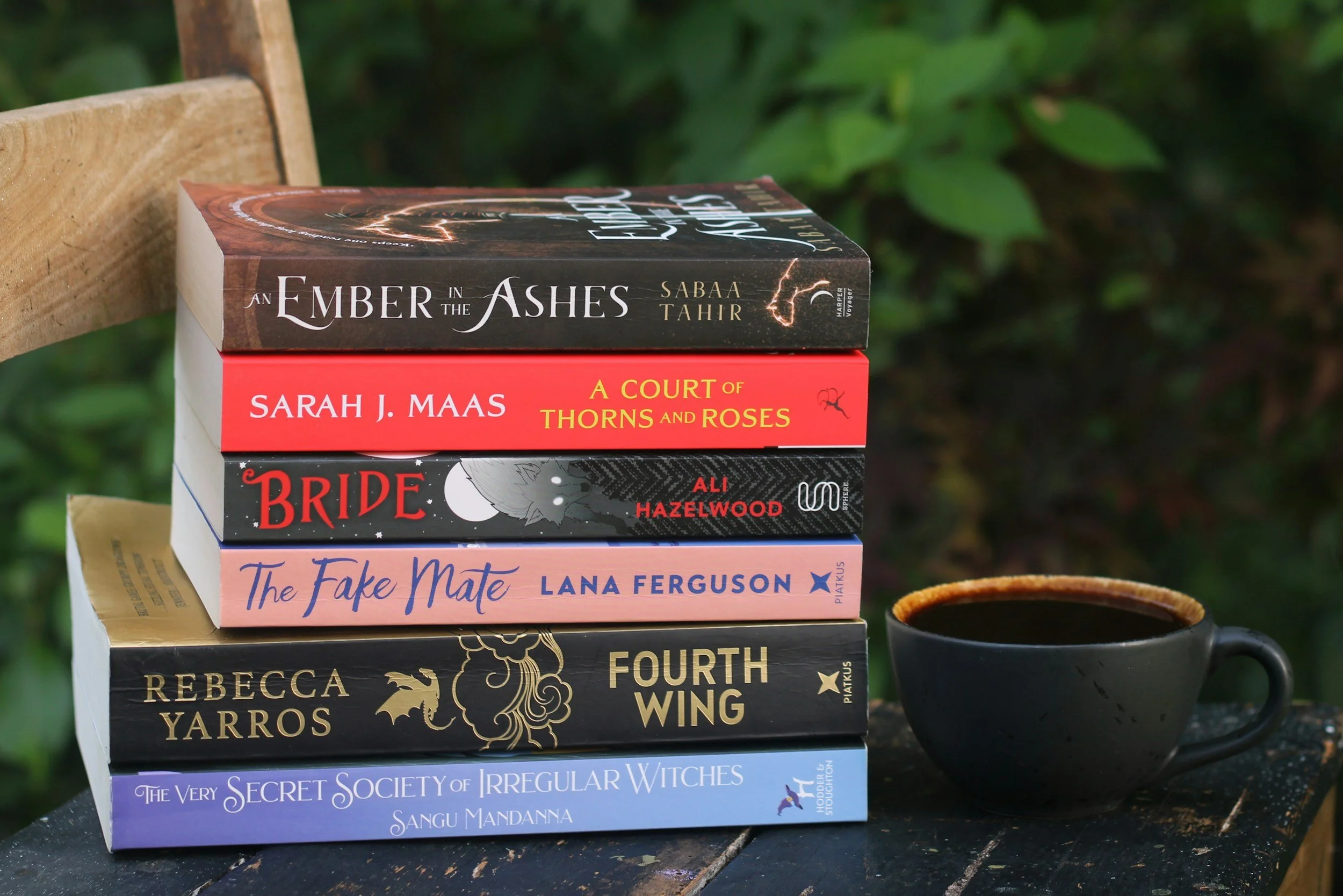 A stack of romantasy books on a dark wooden table with a hot cup of coffee