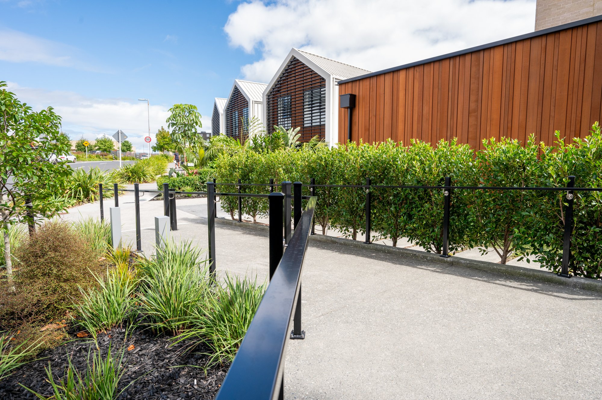 Pathway with black railing and green shrubs, wooden building in background under blue sky.