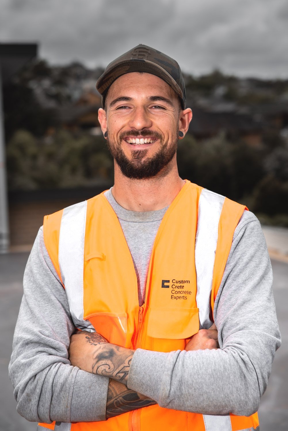 Smiling male construction worker wearing an orange safety vest and cap outdoors with arms crossed.