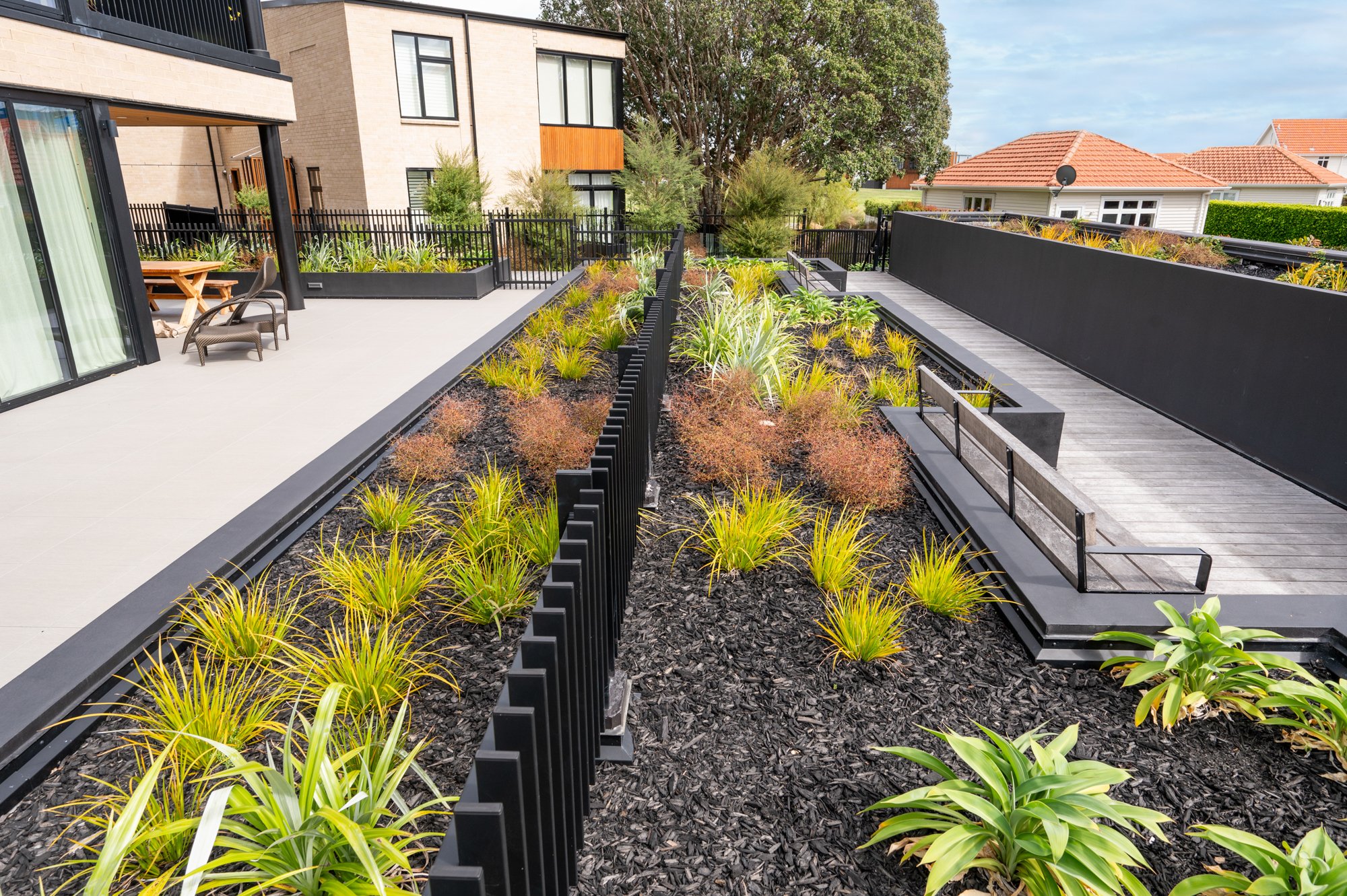 Modern landscaped outdoor area with a paved patio, black fencing, bench seating, garden beds with ornamental grasses, and shrubs. Contemporary house and trees in the background.