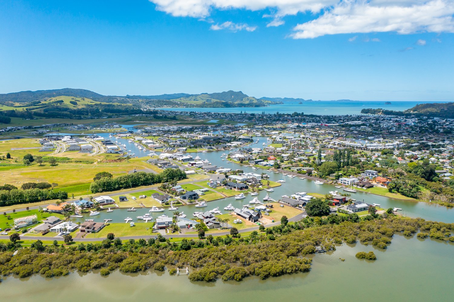 Aerial view of a coastal town with residential areas, waterways, and docks lined with boats, surrounded by green hills and a blue ocean in the background.