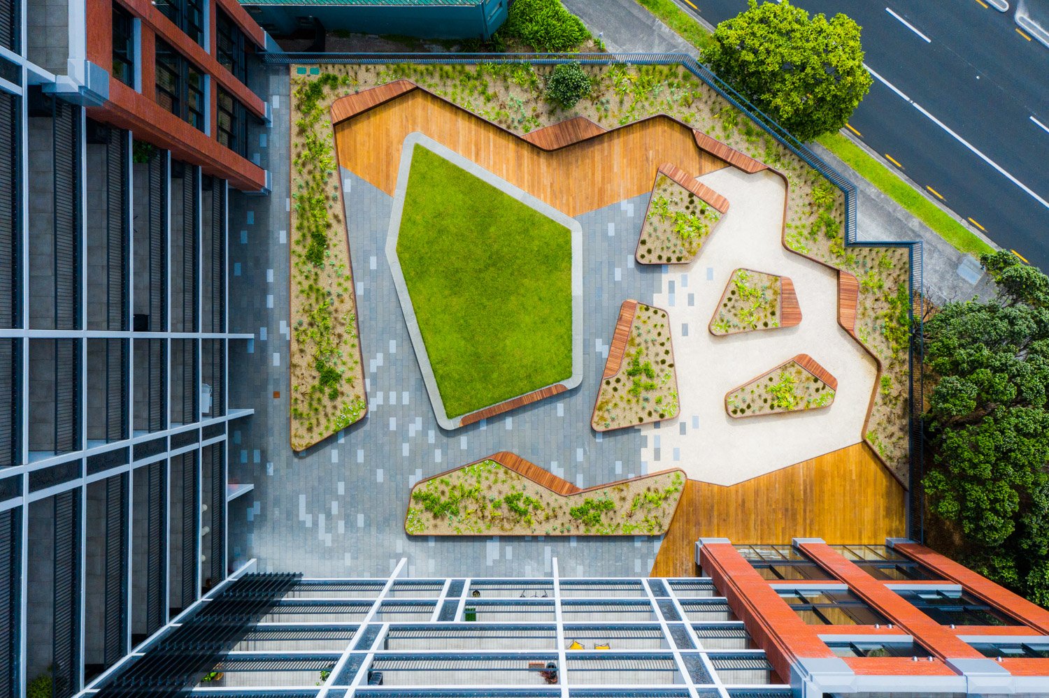 Aerial view of a modern rooftop garden with geometric grass and plant sections, surrounded by pathways and a building with red and black exterior.