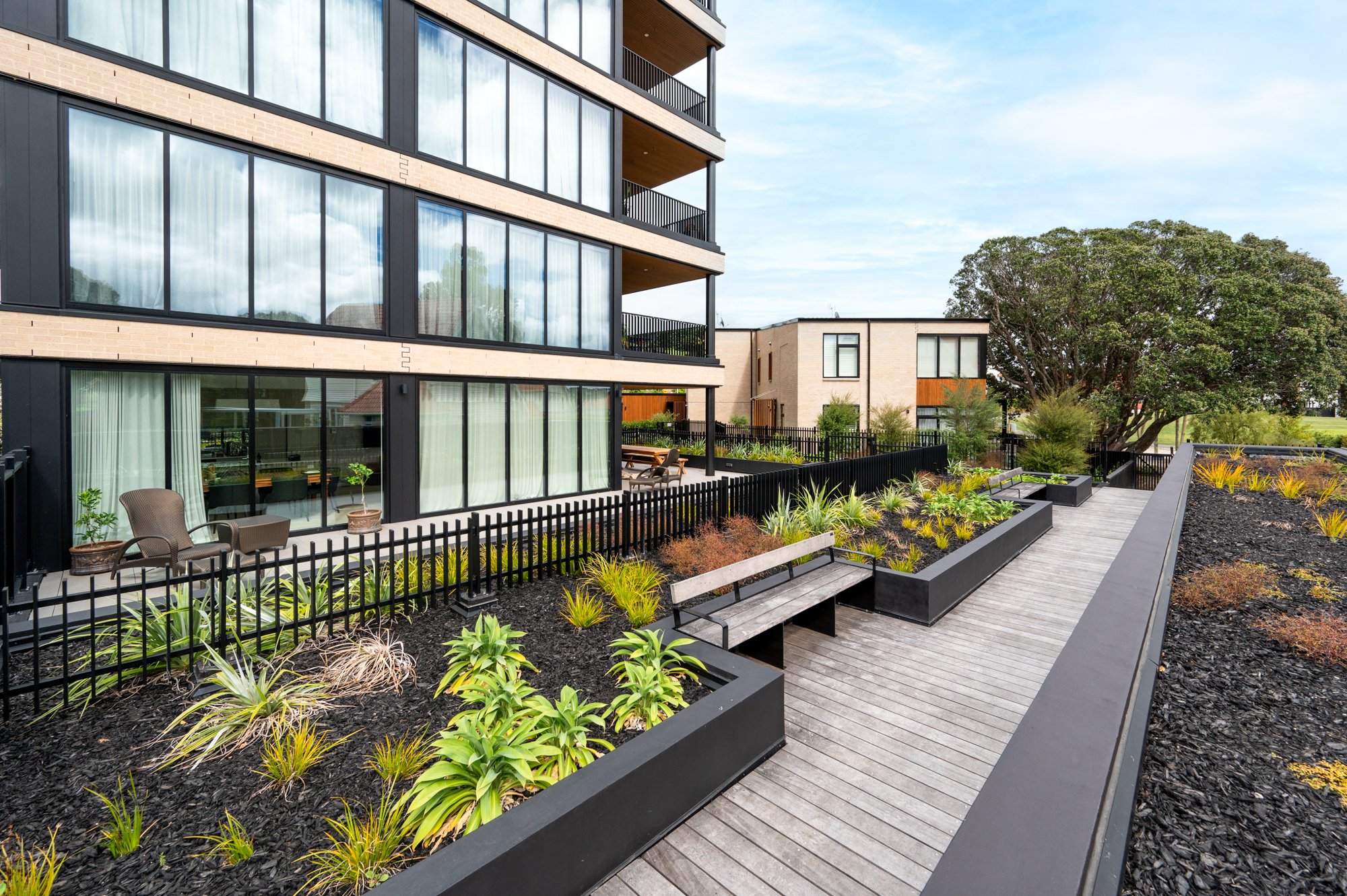 Modern apartment building with large windows, a landscaped courtyard featuring benches and plants, and a wooden pathway.