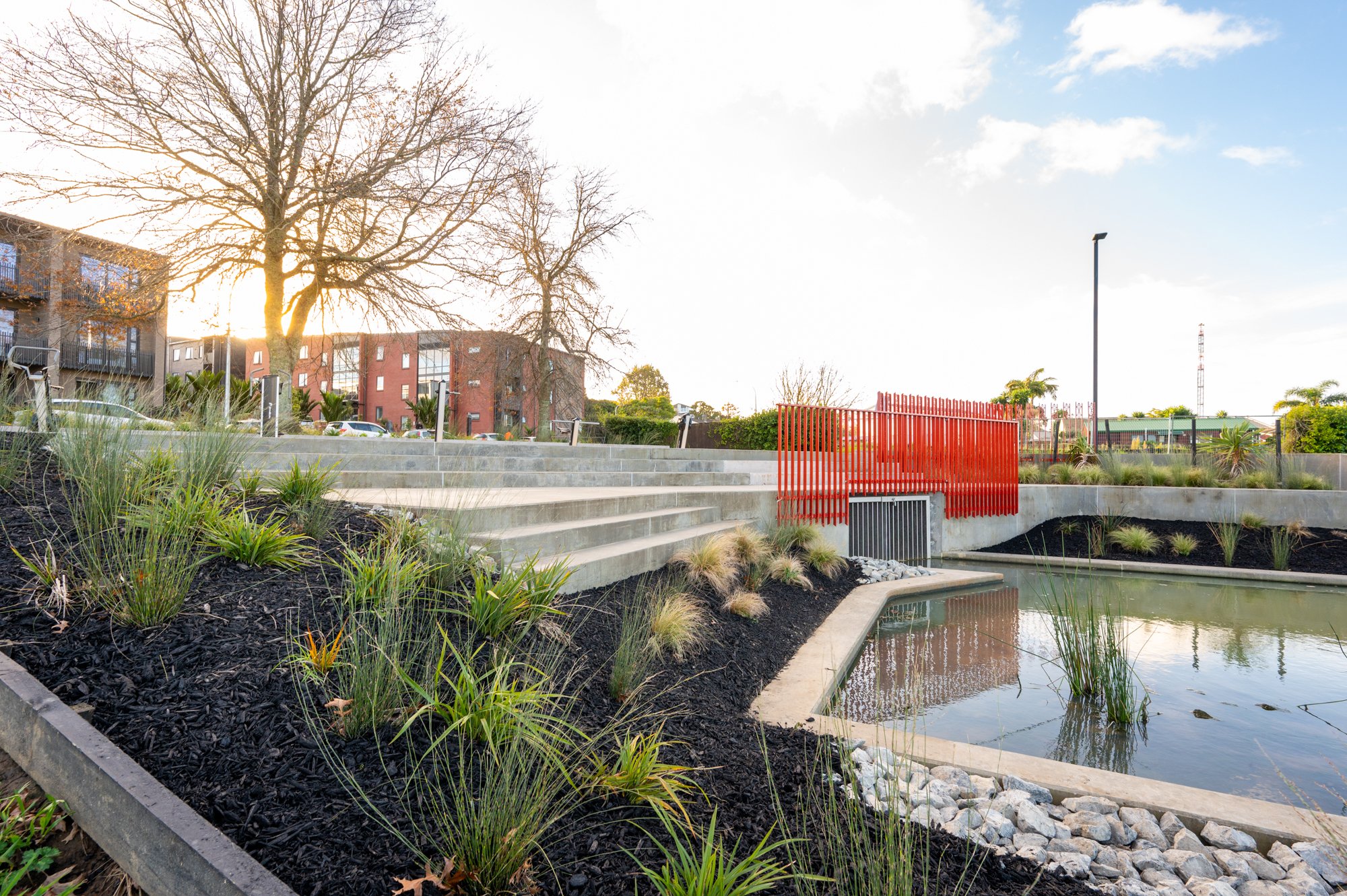 Landscape design with concrete steps, landscaped garden, small pond, and red fence, modern architecture, buildings in background, sunset.
