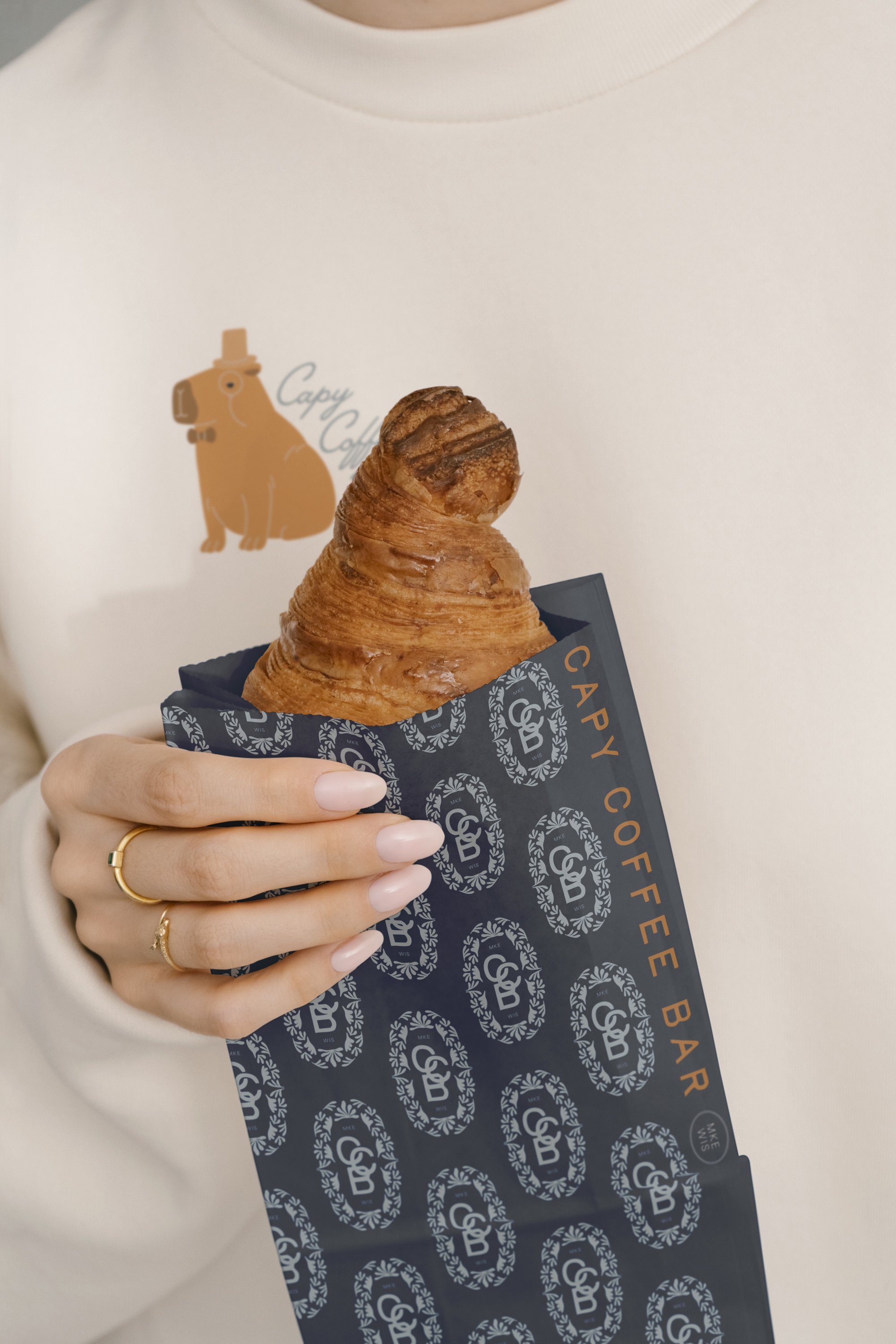 A person holding a croissant in a black branded paper sleeve from Capy Coffee Bar, with a white shirt featuring a dog graphic. The croissant is golden brown and flaky.