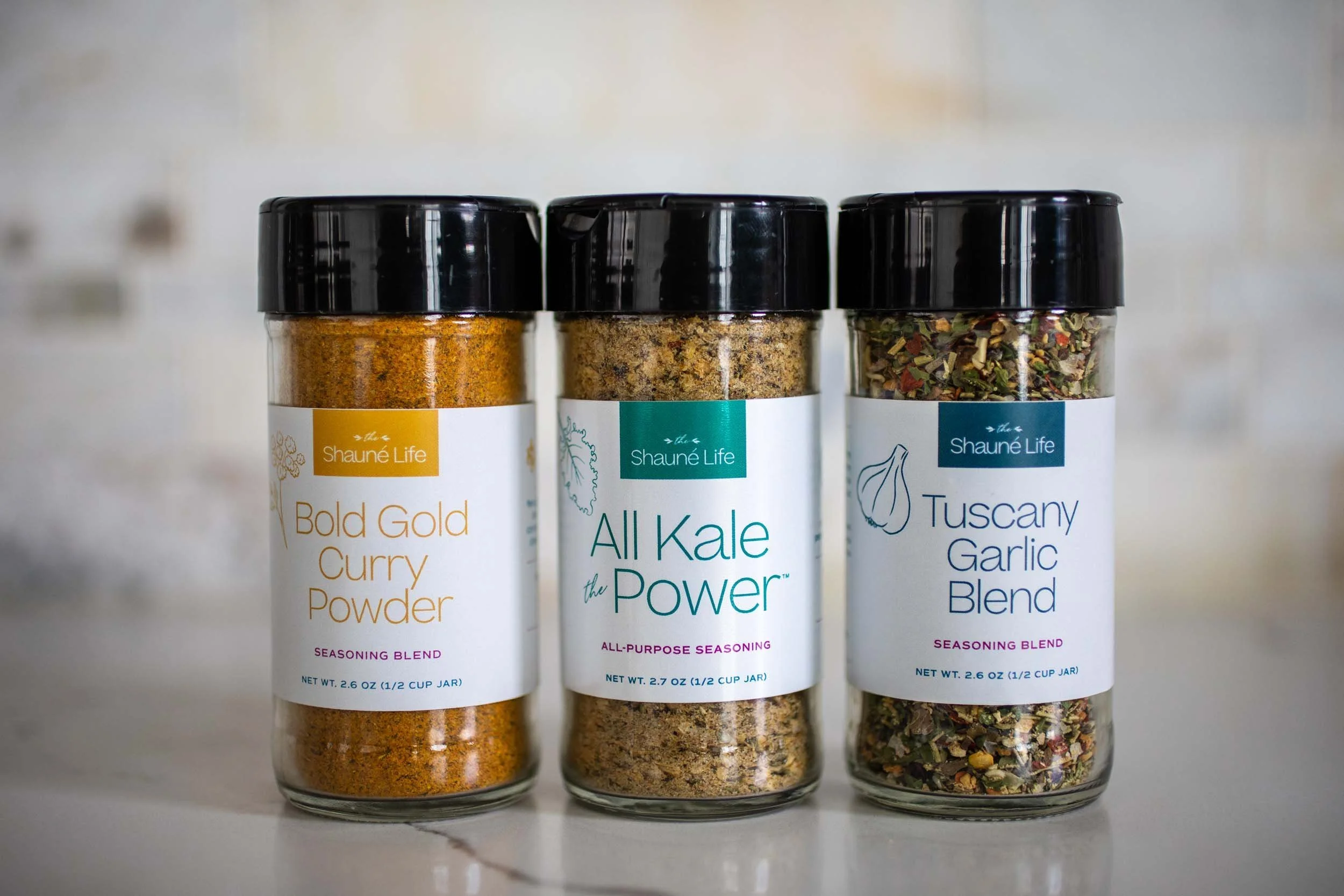 Three glass spice jars with black lids containing curry powder, seasoned kale, and Tuscany garlic blend, each labeled with the brand Shaune Life.