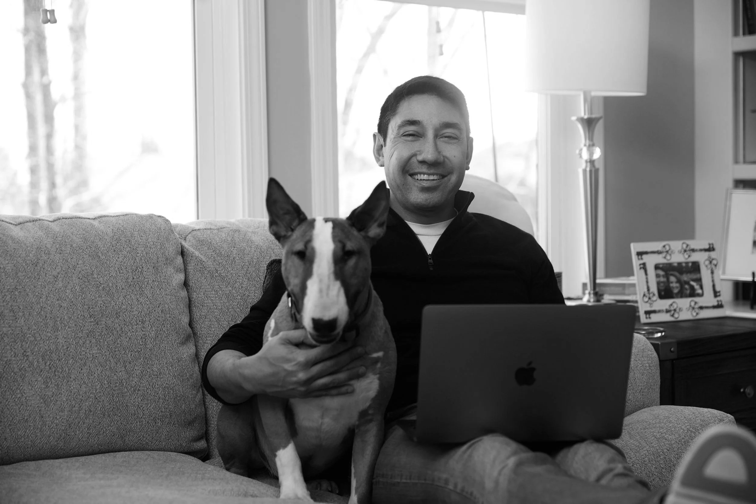 A person sitting on a couch with a laptop and a dog, smiling, in a living room setting.