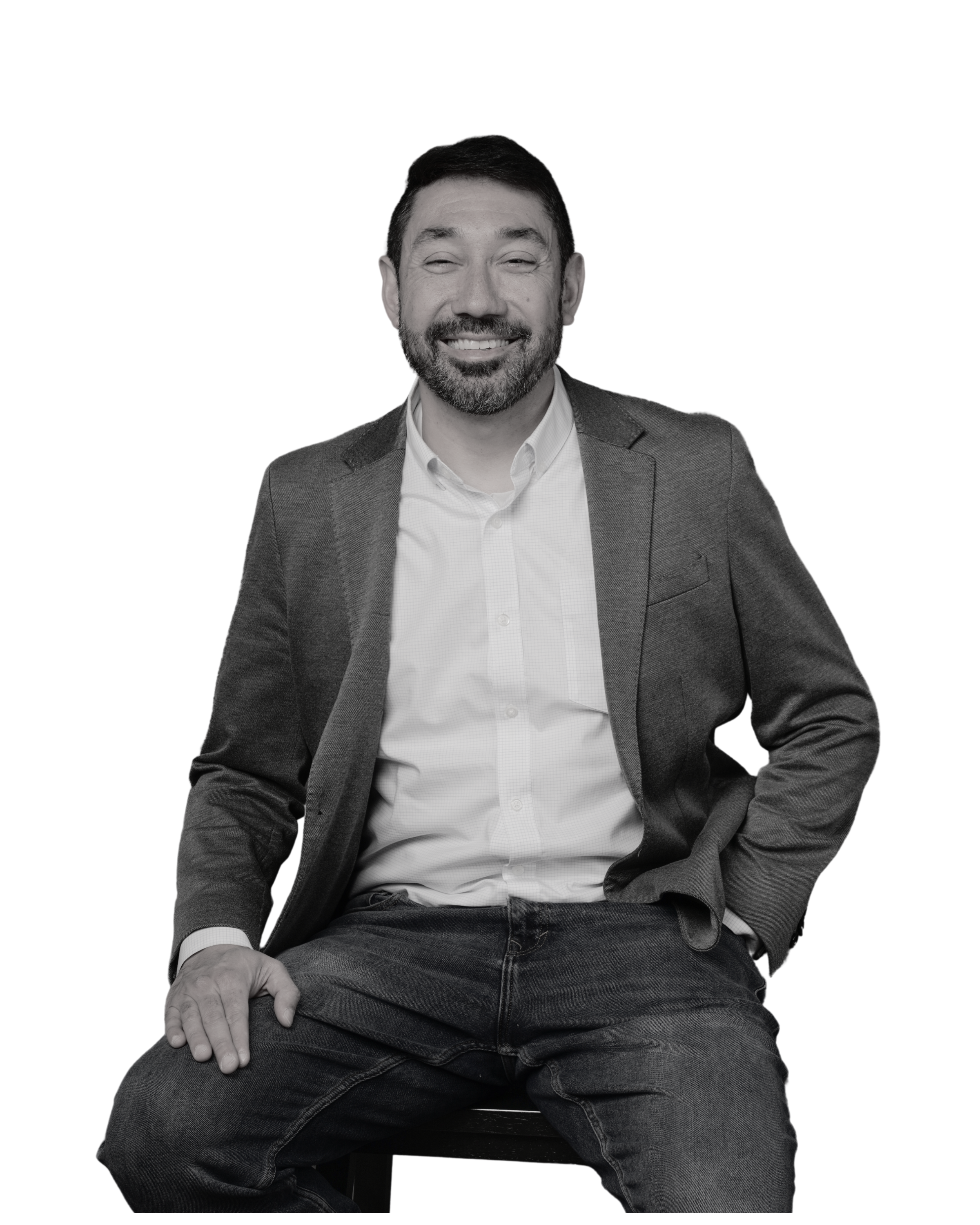 Smiling man sitting in a casual pose, wearing a suit jacket over a shirt, with jeans, and hands resting on his knees.