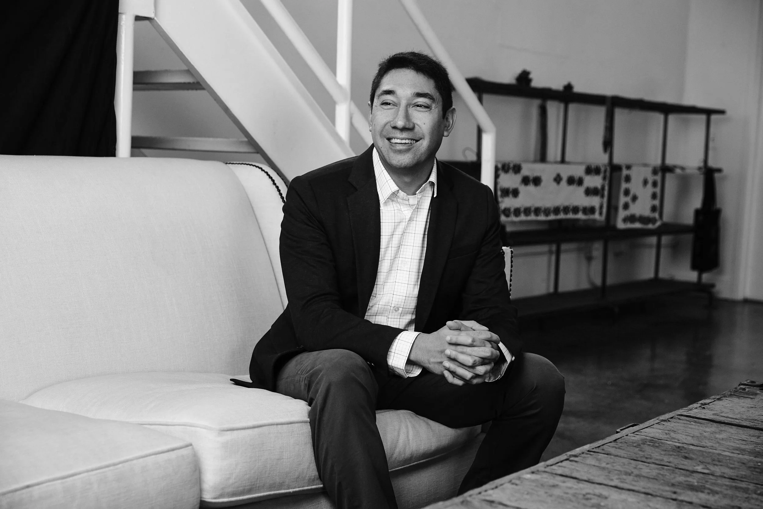 A man in a suit sitting on a couch in a monochrome setting with a staircase and shelves in the background.