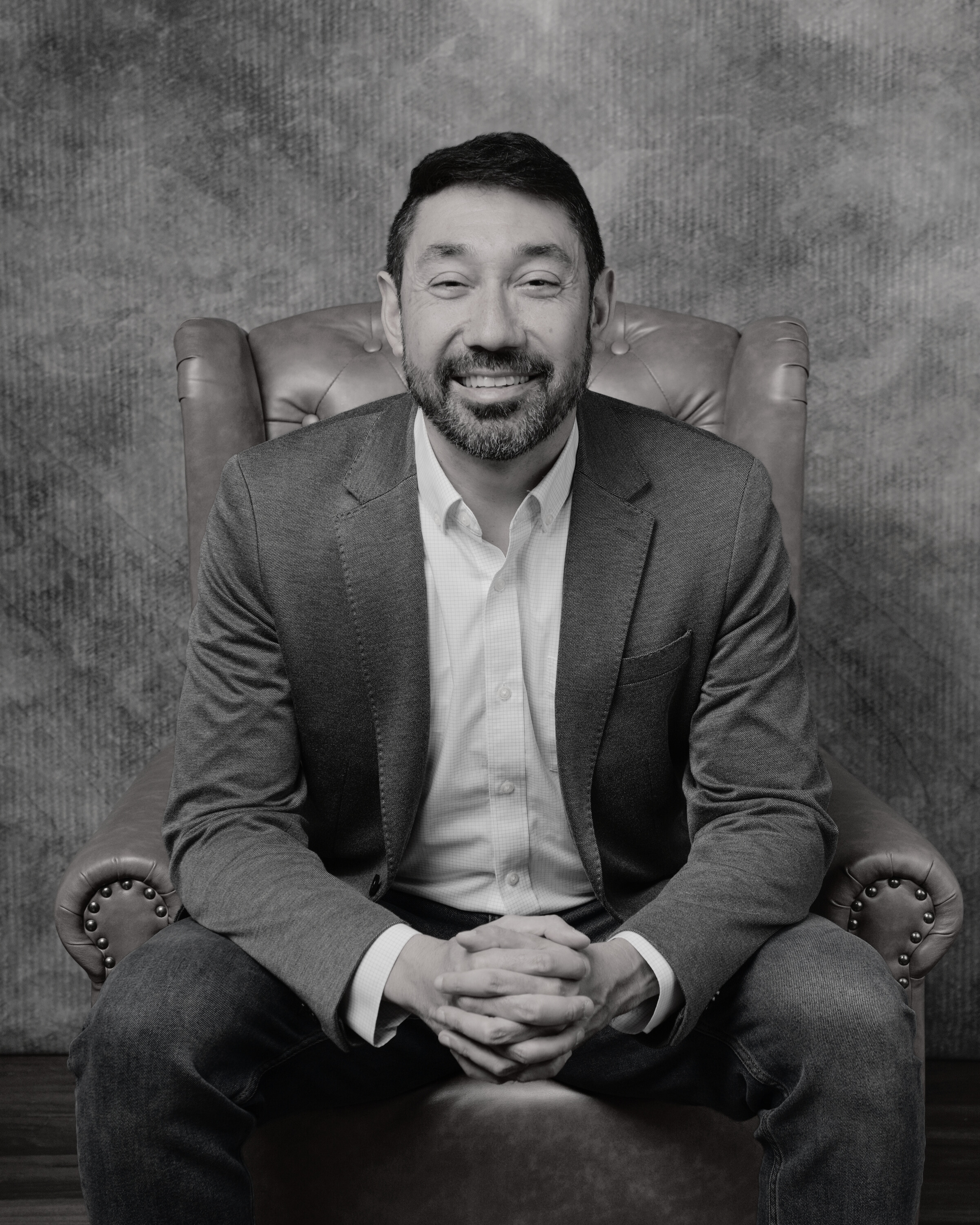 Man sitting in a leather chair, smiling, wearing a suit jacket and shirt, with hands clasped.