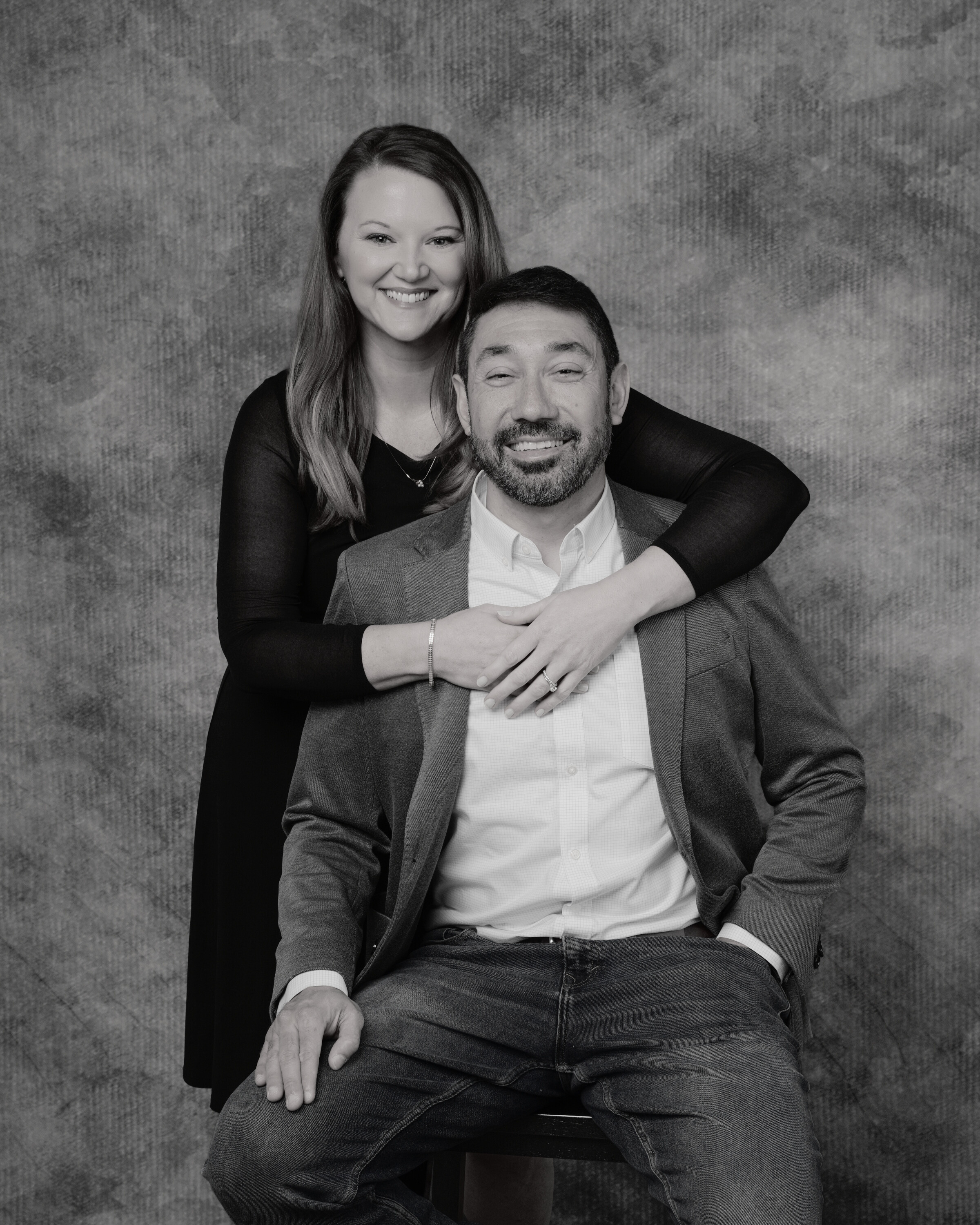 A smiling woman standing behind a seated man with her arms around his shoulders, both dressed casually.