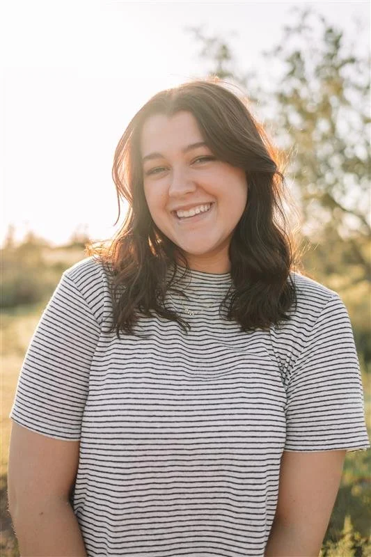 A smiling young woman with shoulder-length dark hair, wearing a striped t-shirt, standing outdoors in natural light with trees in the background.