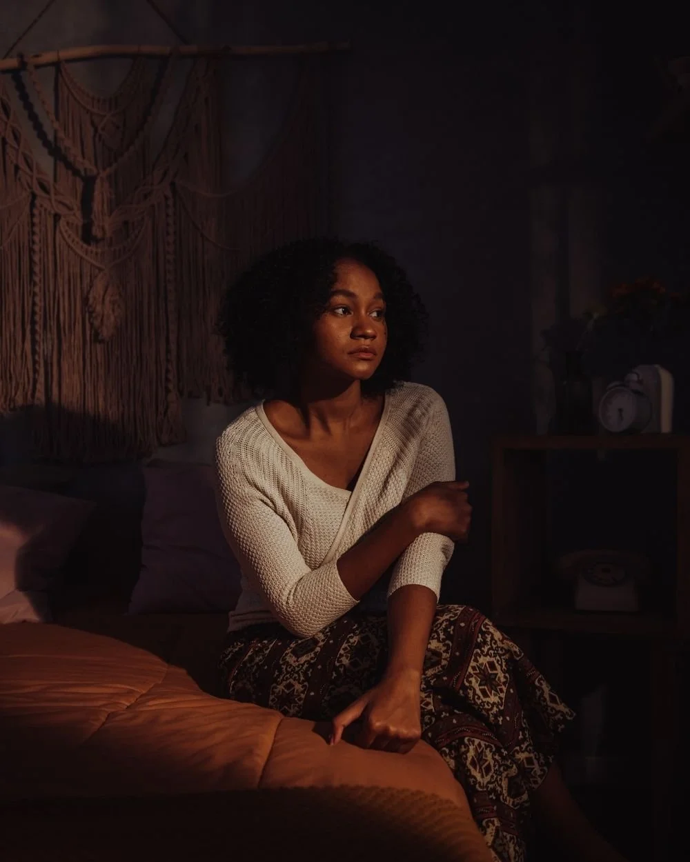 A young Black woman sitting alone on her bed in soft lamplight, looking away in thought.