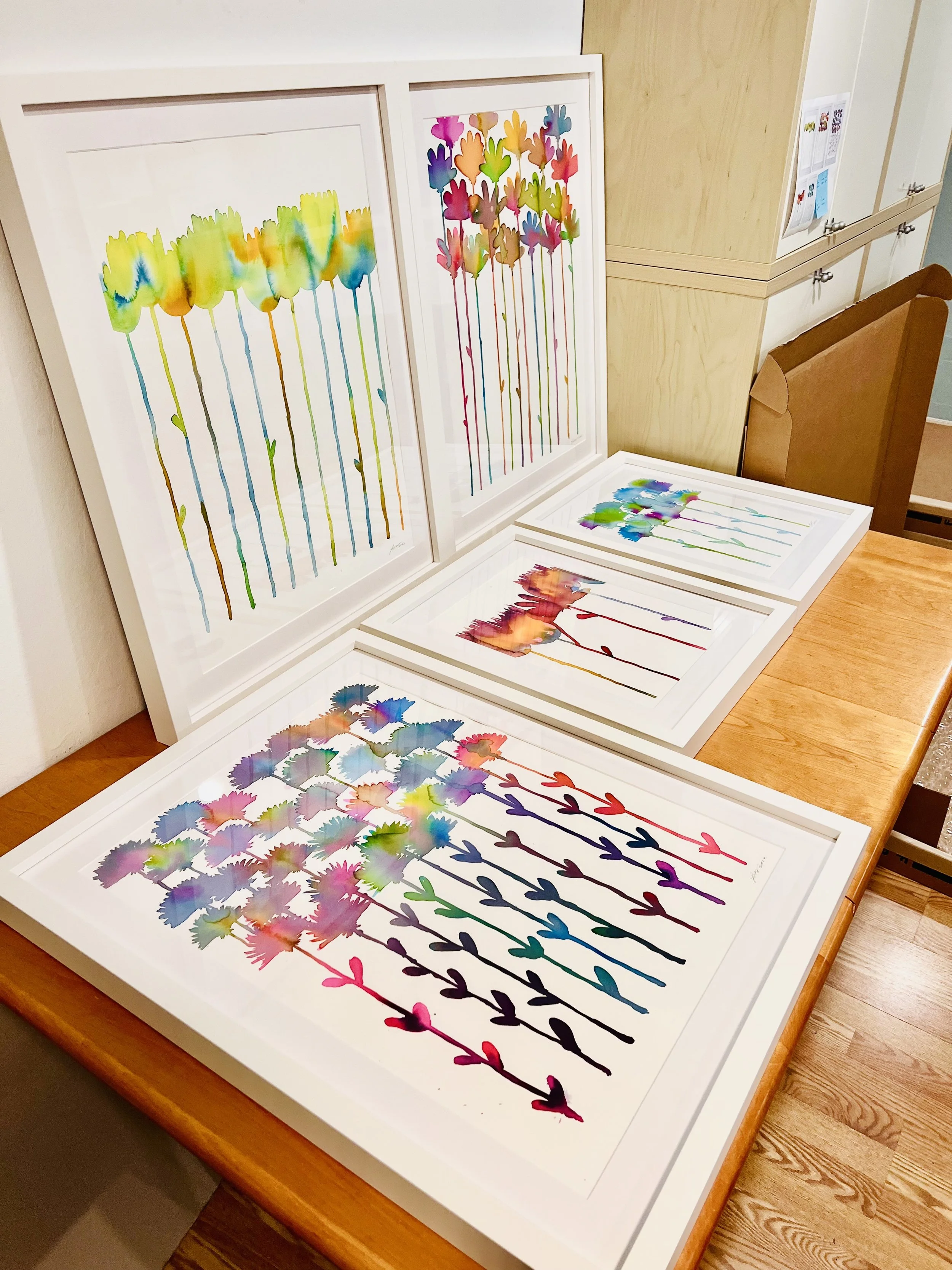 Colorful watercolor paintings of flowers on a wooden table, featuring abstract, vibrant flower patterns with drips and splashes.