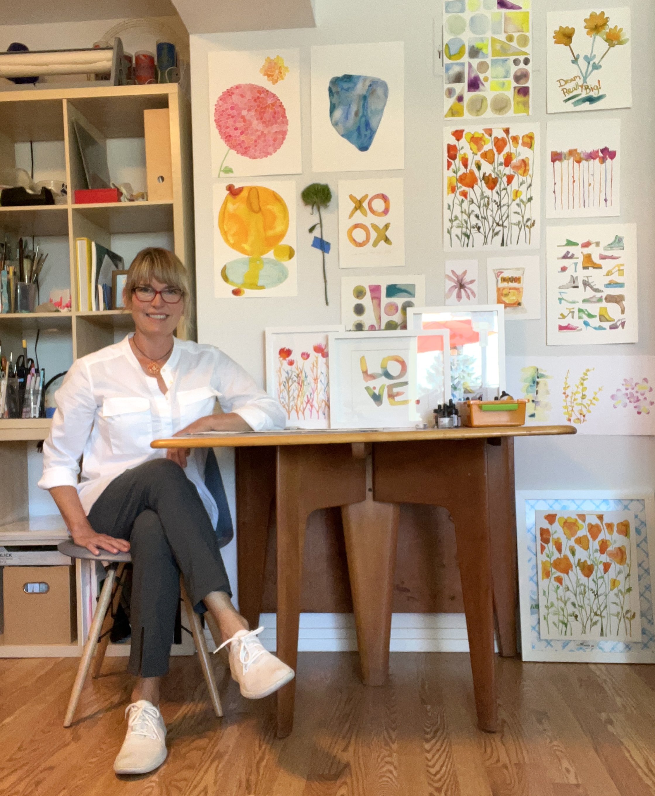 A woman sitting at a wooden table in an art studio, surrounded by colorful watercolor paintings of flowers, rocks, and abstract designs on the wall behind her.