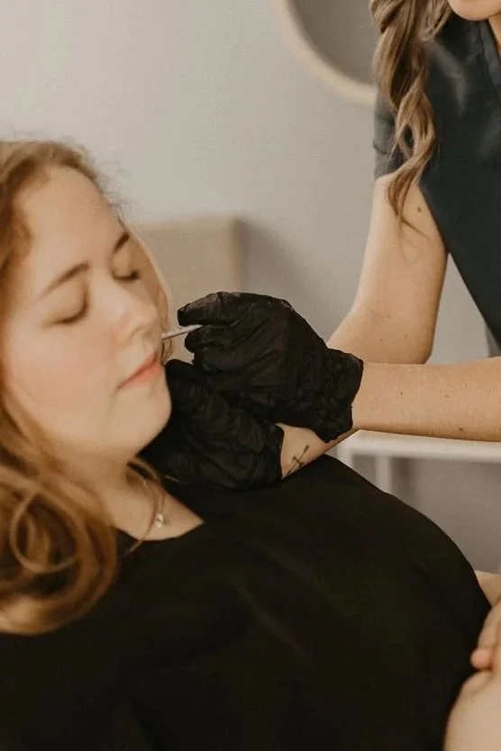 A woman in medical scrubs performing a facial procedure on a reclining patient in a clinical setting.