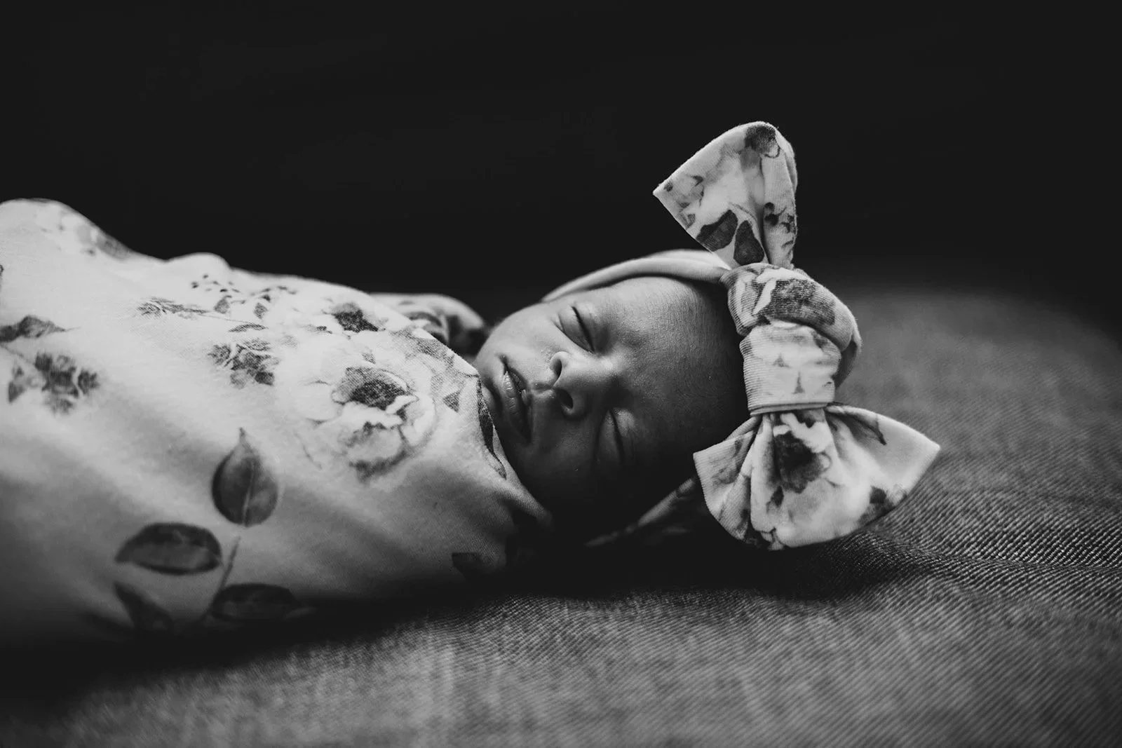 Black and white photo of a sleeping baby wrapped in a floral blanket with a matching bow on their head.
