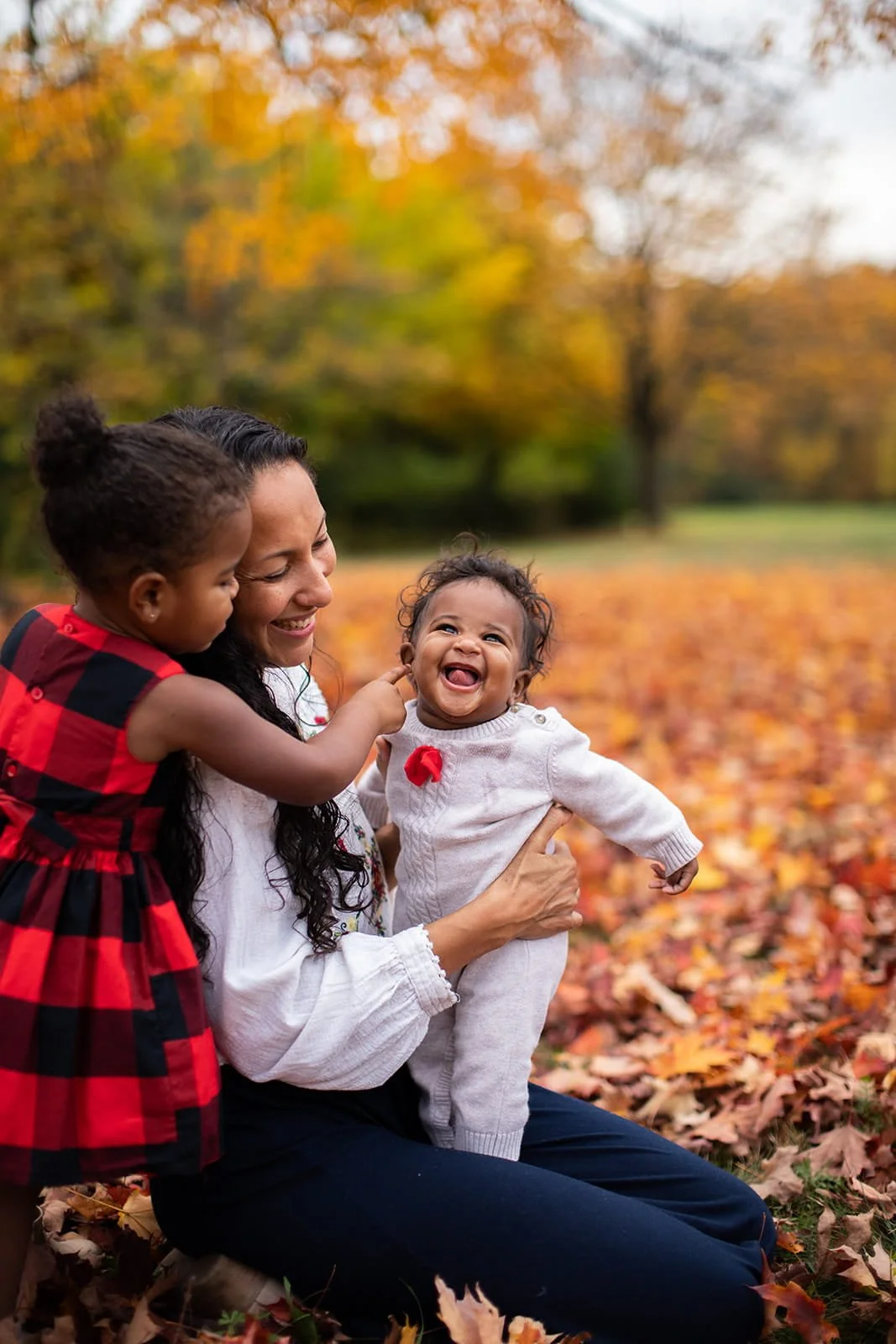 A woman with two young girls in an autumn park with colorful fallen leaves and trees with orange and yellow foliage. The woman is smiling and holding a toddler girl in a white sweater, while another young girl in a red and black plaid dress is touching the toddler's face.