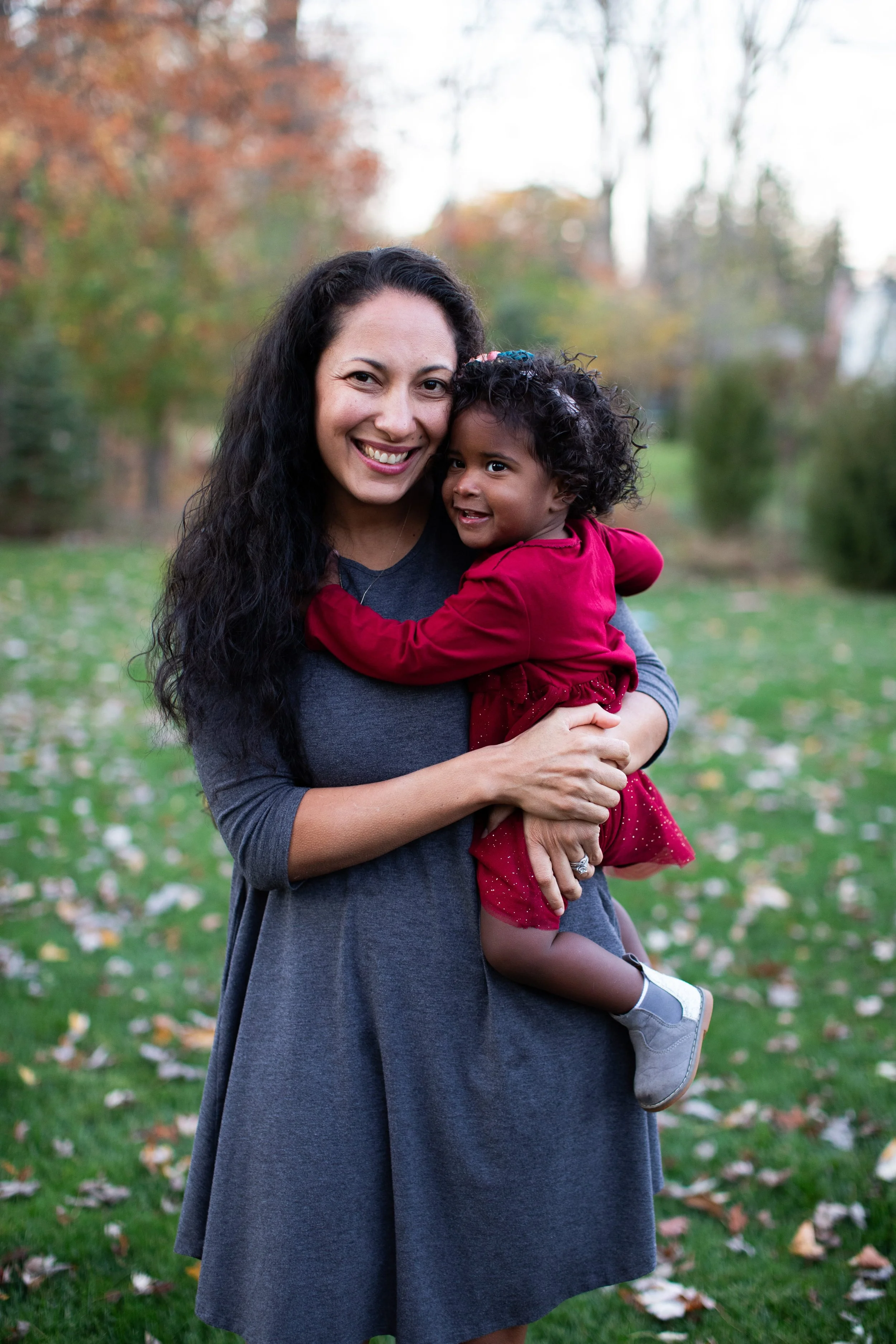 A woman with long dark hair smiling and holding a young girl with curly hair, dressed in red, during autumn in a park with fallen leaves and trees in the background.