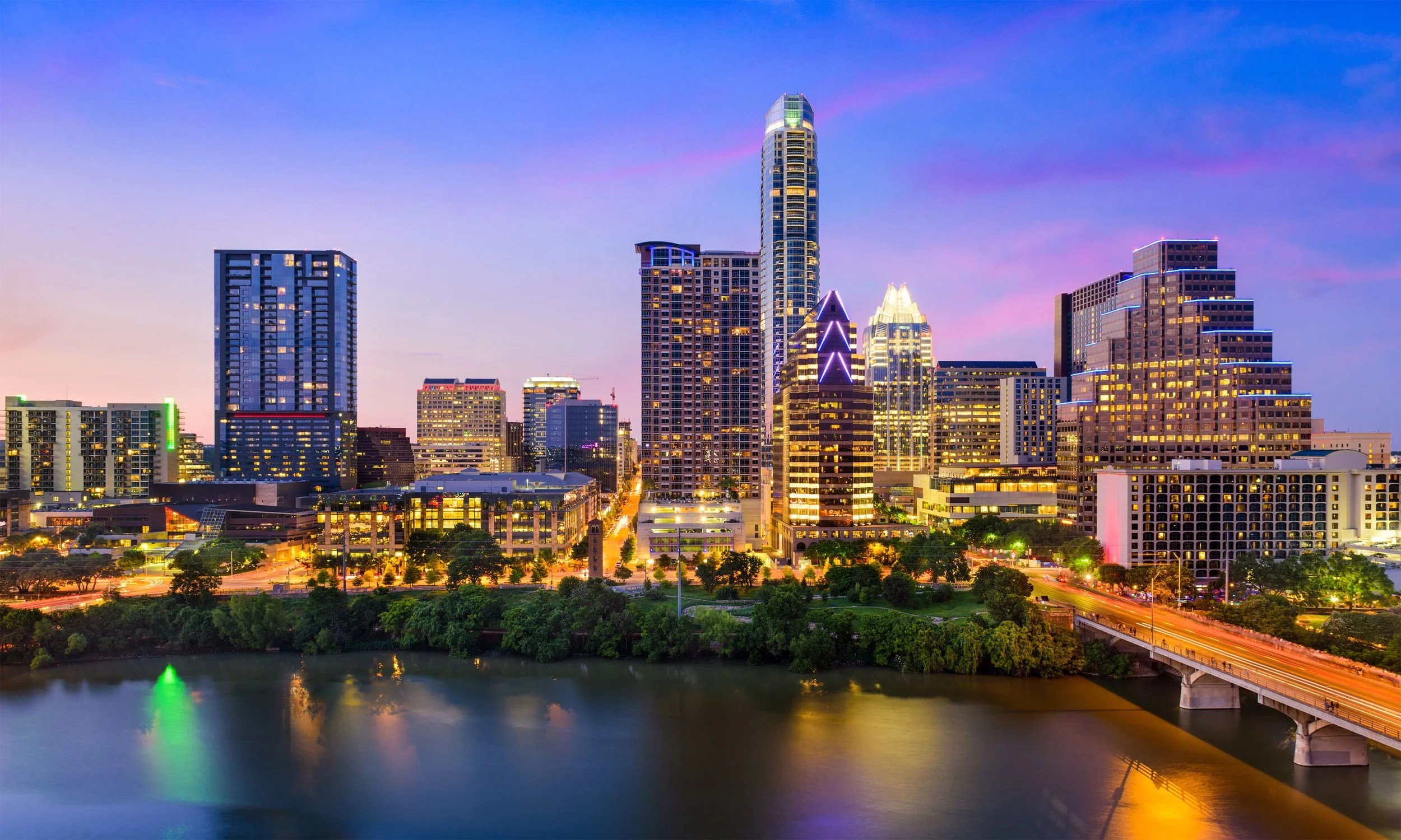 Night view of a city skyline with illuminated skyscrapers, a river in the foreground, and a bridge with traffic.