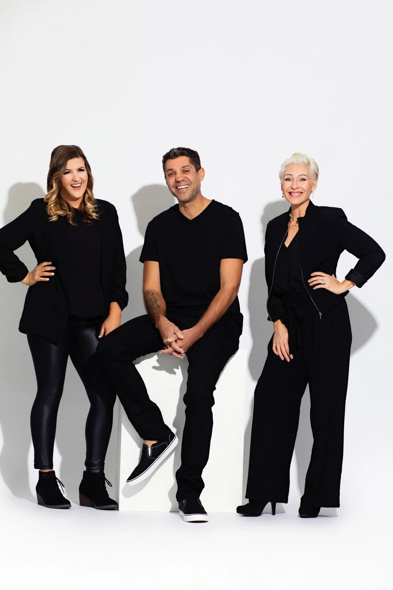 Three adults standing against a white background, dressed in black, smiling and posing for the camera.