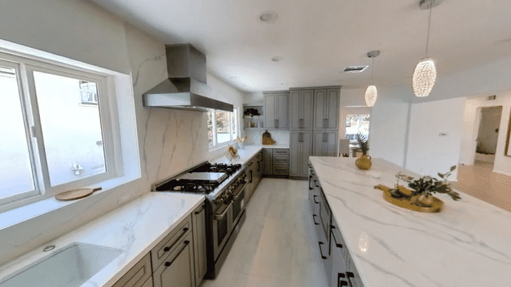 Modern kitchen with white marble countertops, gray cabinets, and stainless steel appliances. Two pendant lights hang above the island, and there are large windows letting in natural light.