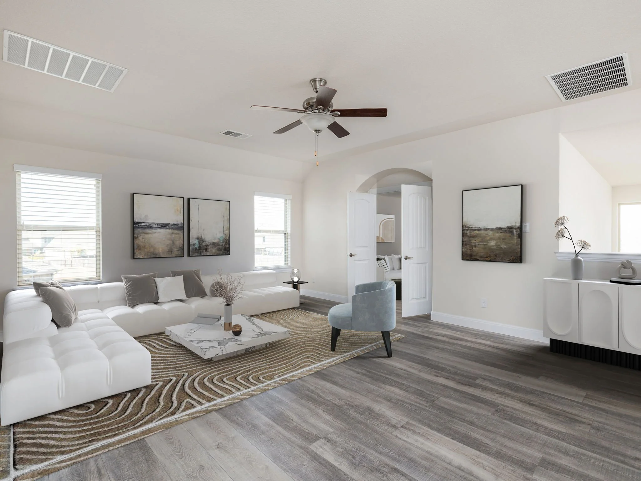 Bright living room with white walls, a large white sectional sofa with pillows, a marble coffee table, two paintings on the wall, small windows with blinds, a ceiling fan, a blue accent chair, and a sideboard with decorative items.