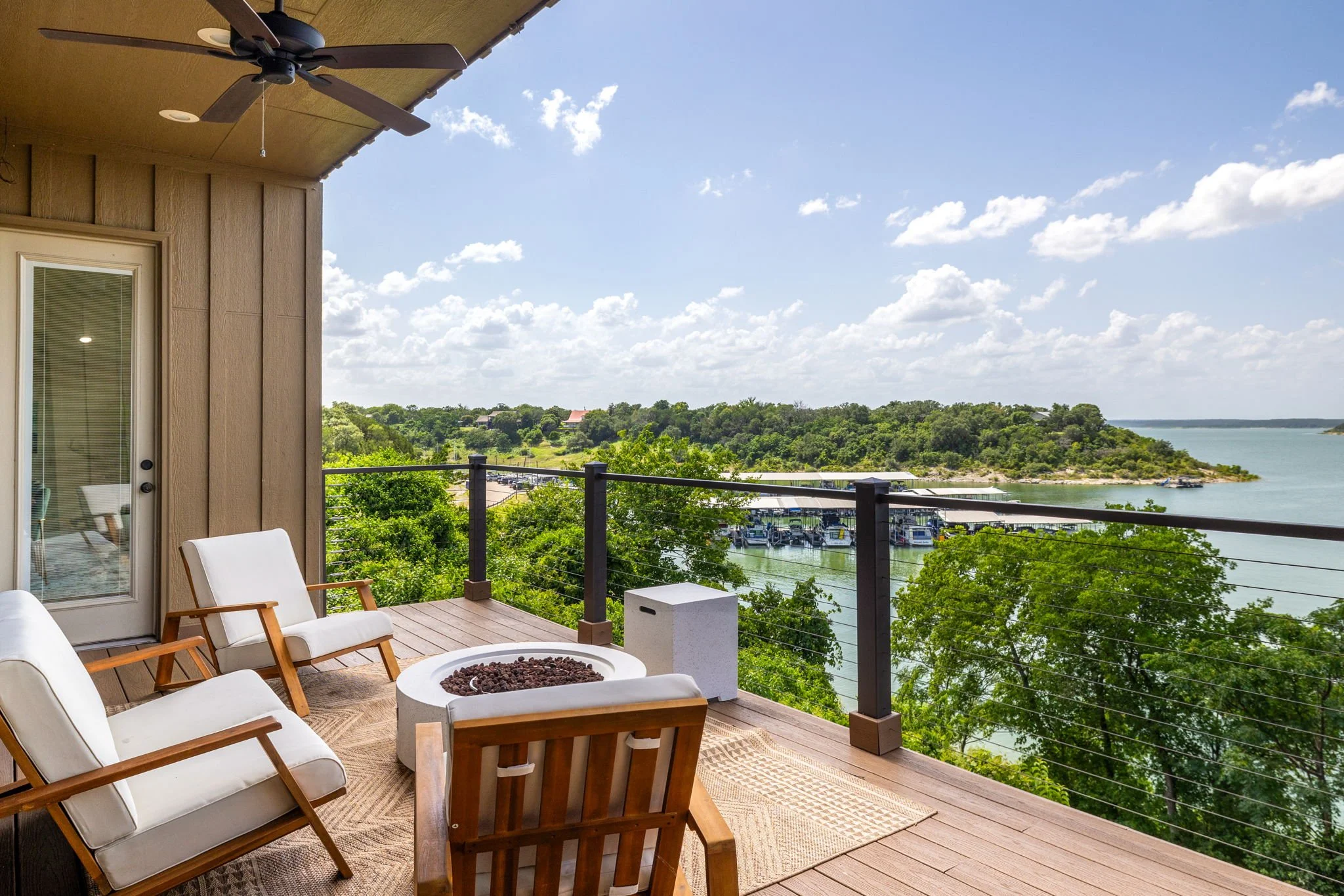 Balcony with outdoor furniture including three wooden chairs with white cushions around a fire pit, overlooking green trees, a marina, and a lake under a partly cloudy sky.