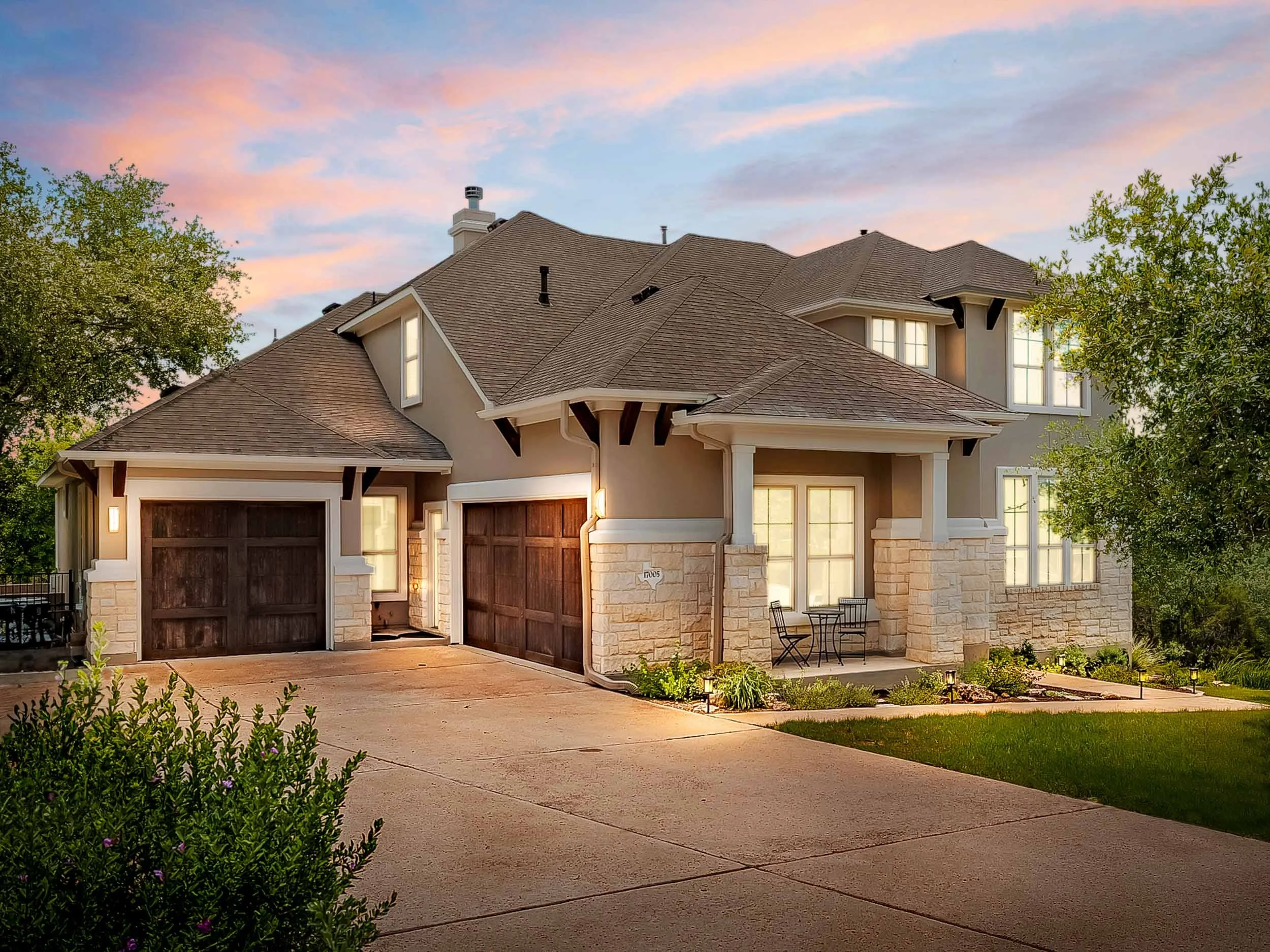 A modern two-story house with stone accents, wooden garage doors, and a landscaped front yard during dusk with a colorful sky.