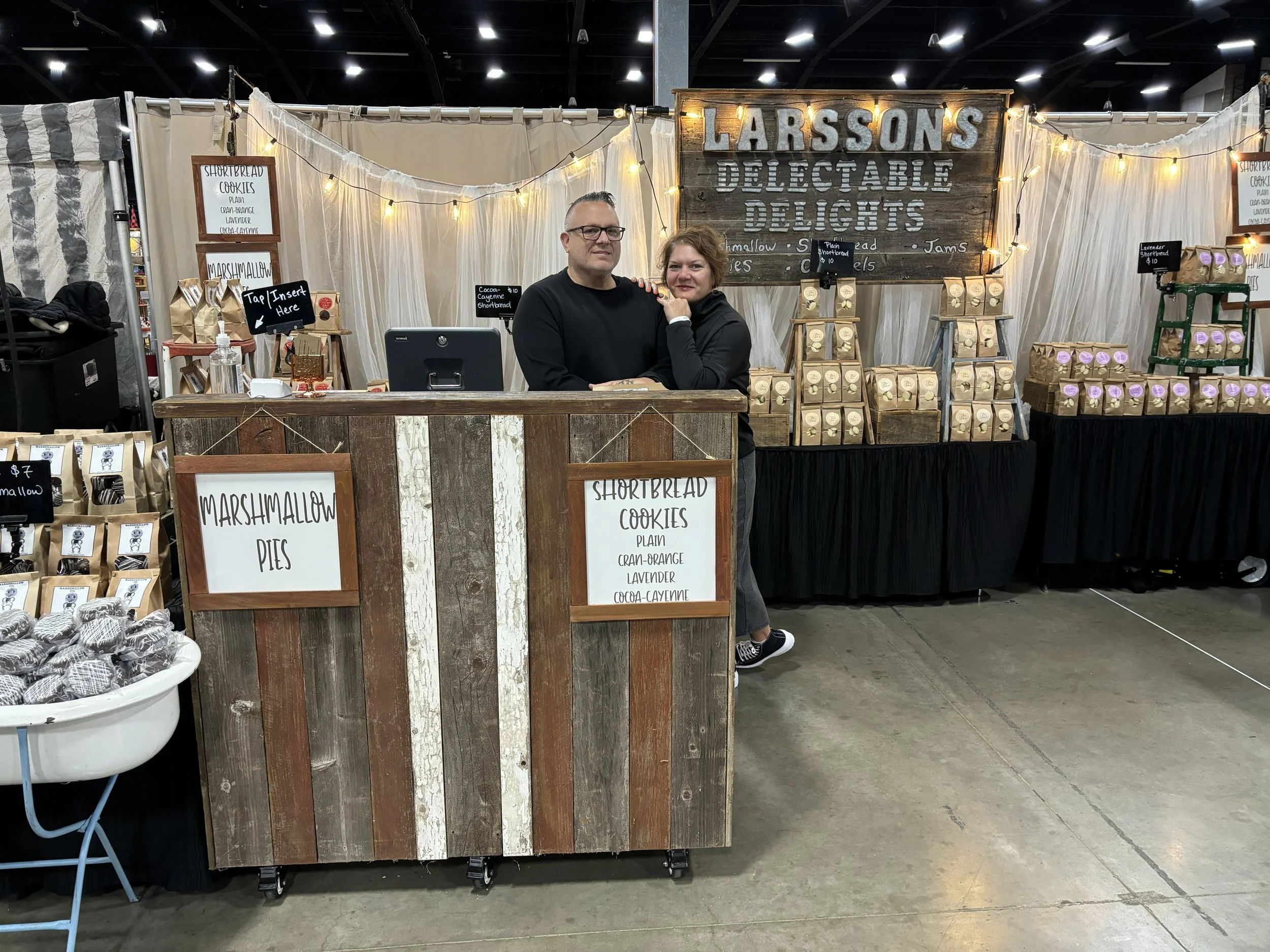 Rob and Lorraine Larsson standing behind a rustic wooden booth at a market, with signs advertising marshmallow pies and shortbread cookies, and a large sign reading 'LARSSON'S DELICATABLE DELIGHTS' in the background.