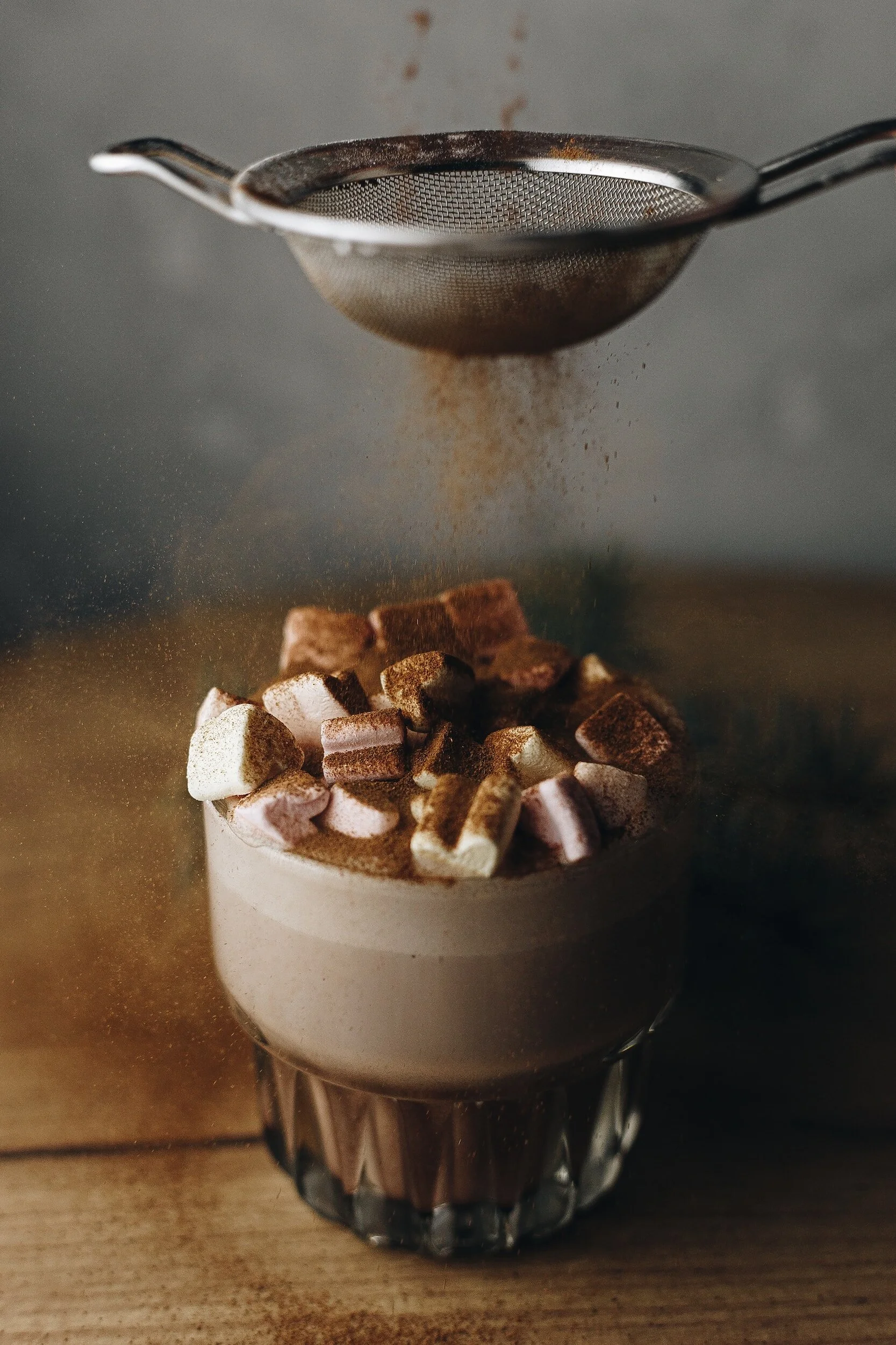 Marshmallows being dusted with cocoa powder over a layered beverage in a glass on a wooden surface.