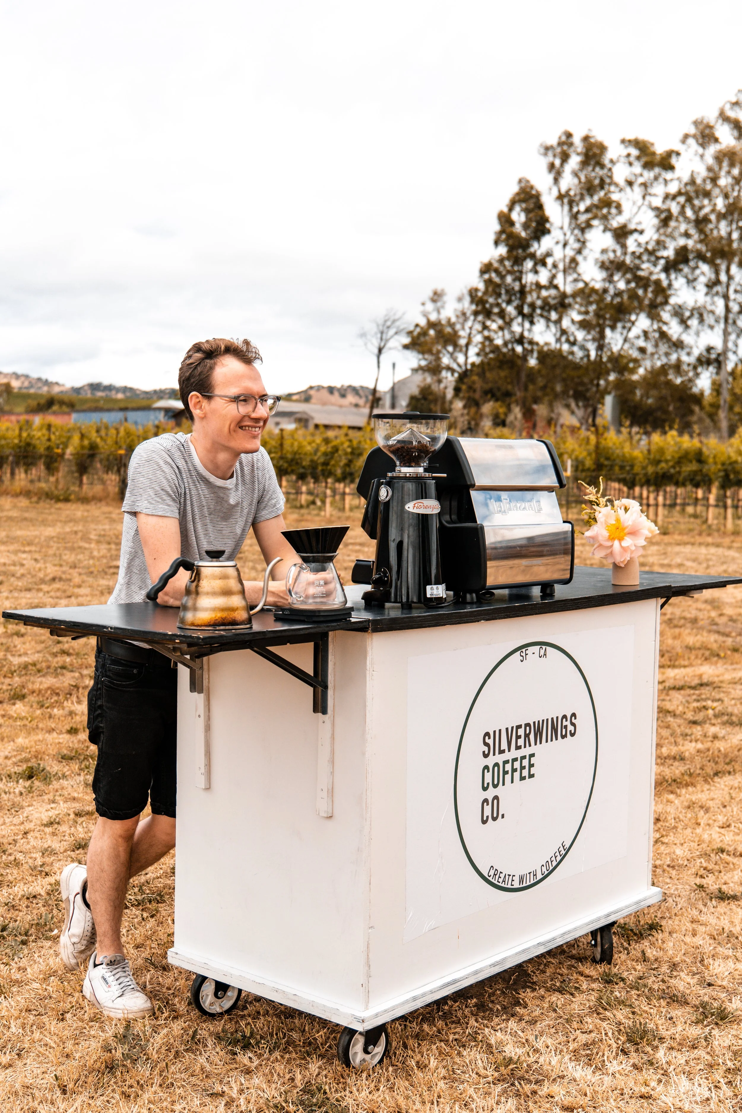 A man smiling at a coffee stand on a grassy field, with trees and vineyards in the background, serving coffee from a black espresso machine labeled 'Silverwings Coffee Co.'