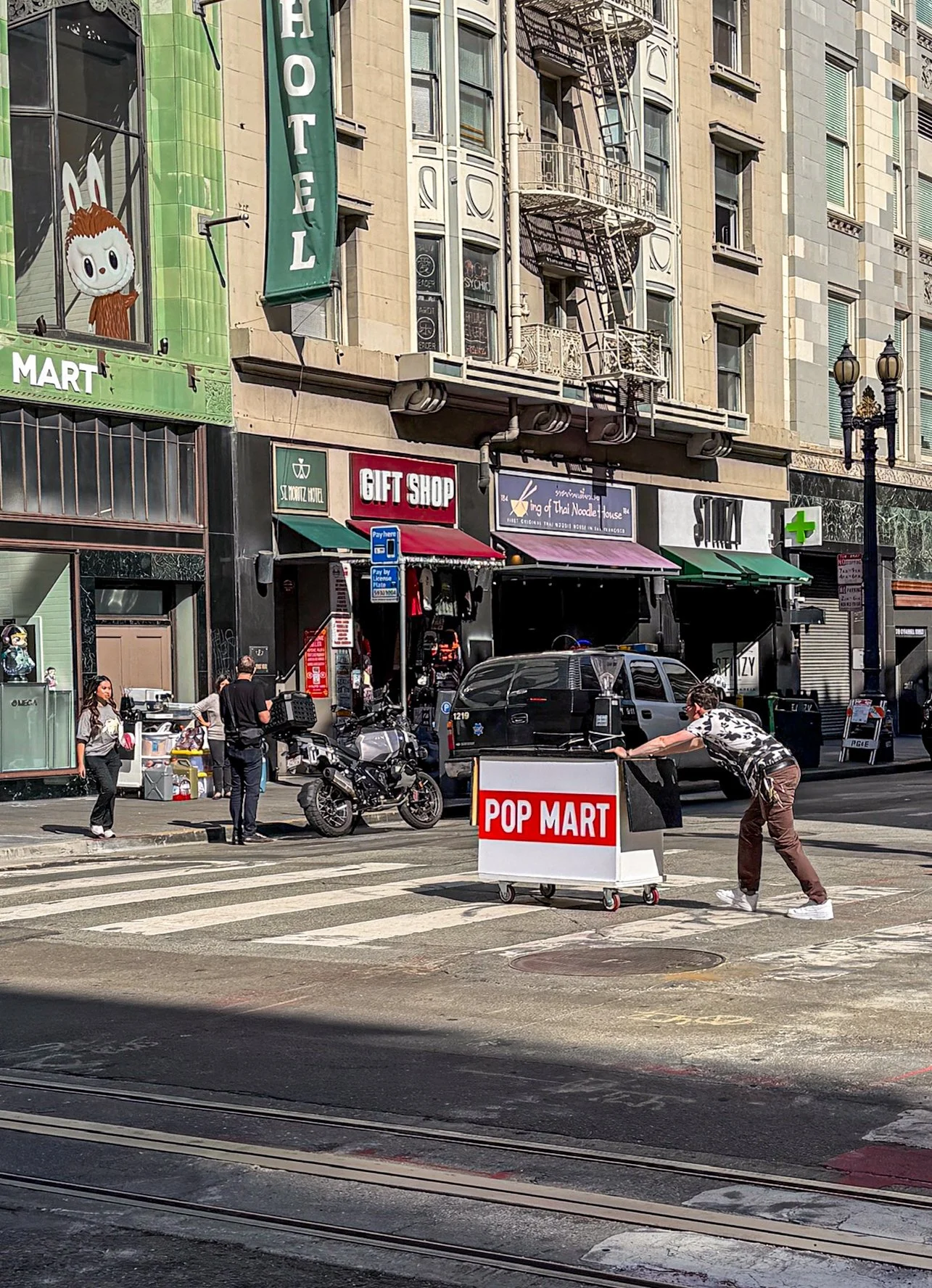 City street scene with pedestrians crossing, a person pushing a cart labeled 'POP MART,' storefronts including a gift shop and Thai noodle restaurant, parked motorcycle, and an apartment building with fire escapes.