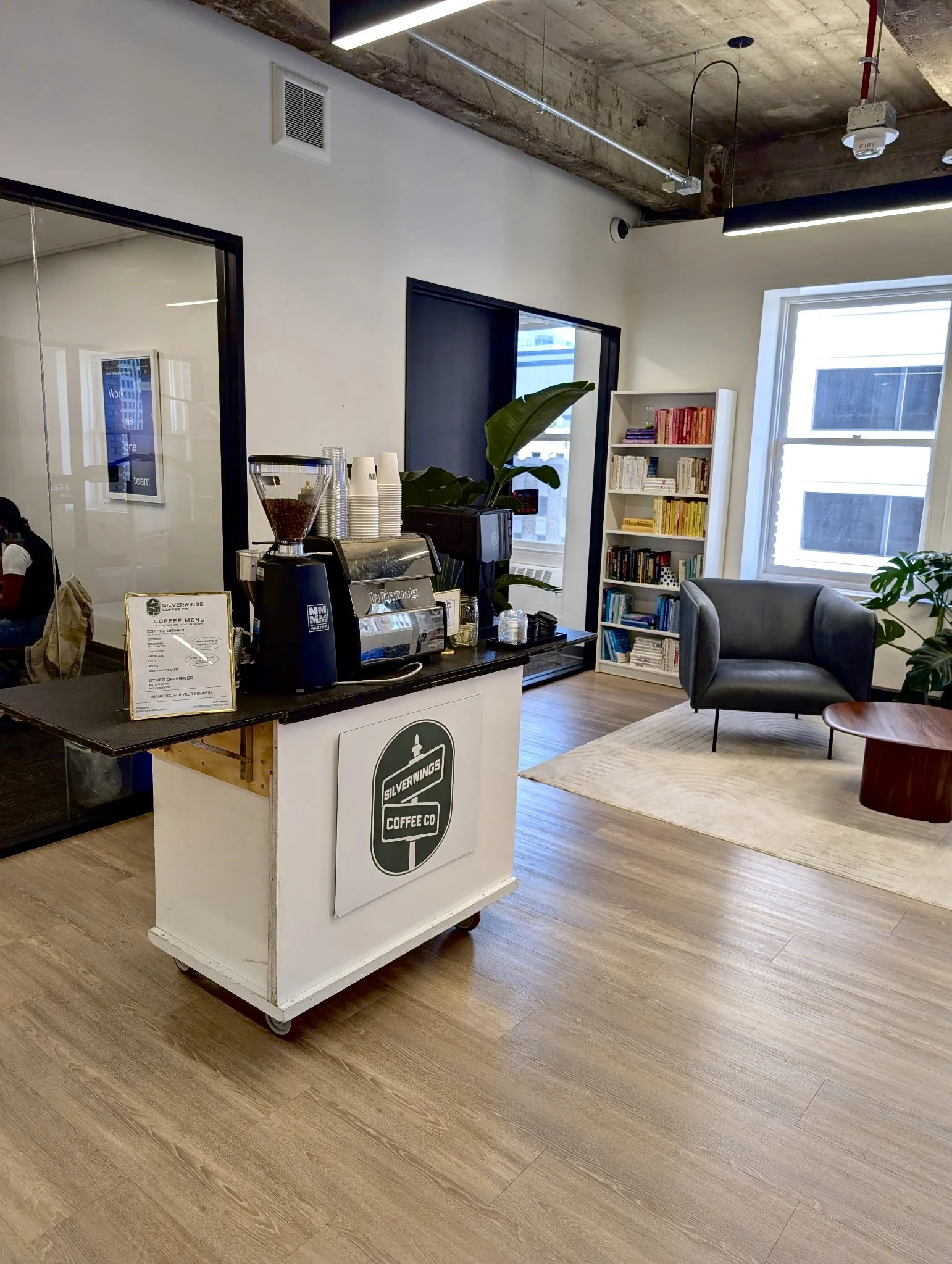 Modern office with mobile coffee cart with an espresso machine, cups, and a menu on it. There is a large plant, a meeting room and bookshelves in the background, with wooden floors, concrete ceiling, and large windows letting in natural light.