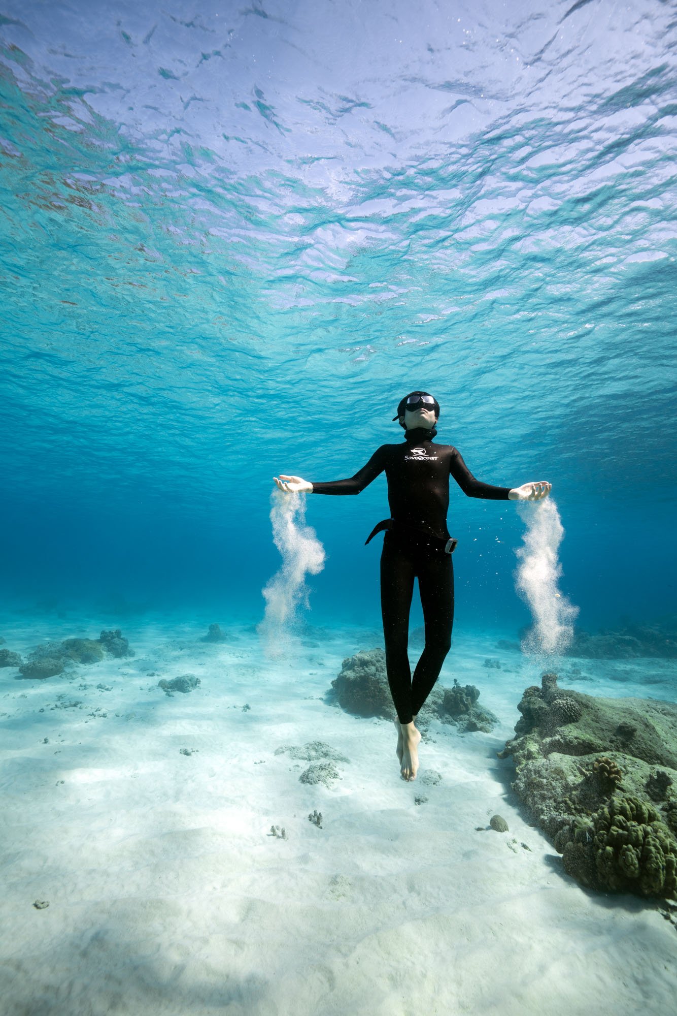 Freediver underwater during a breath-hold dive, letting sand flow through their fingers above a sandy ocean seabed.