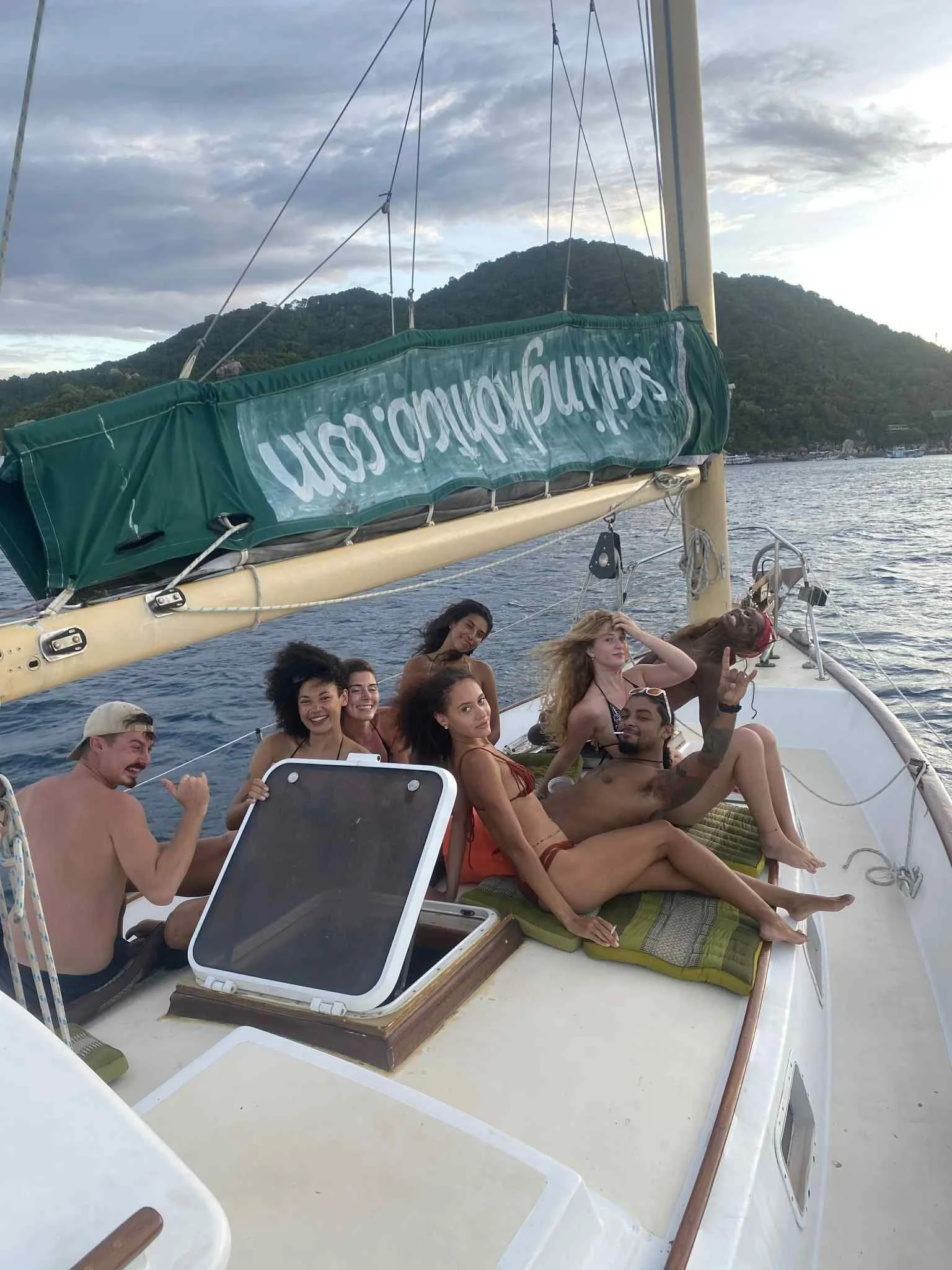 Guests relaxing and smiling onboard Orenda Freediving & Island Cruises sailboat with Koh Tao island and blue sky in background during sailing cruise