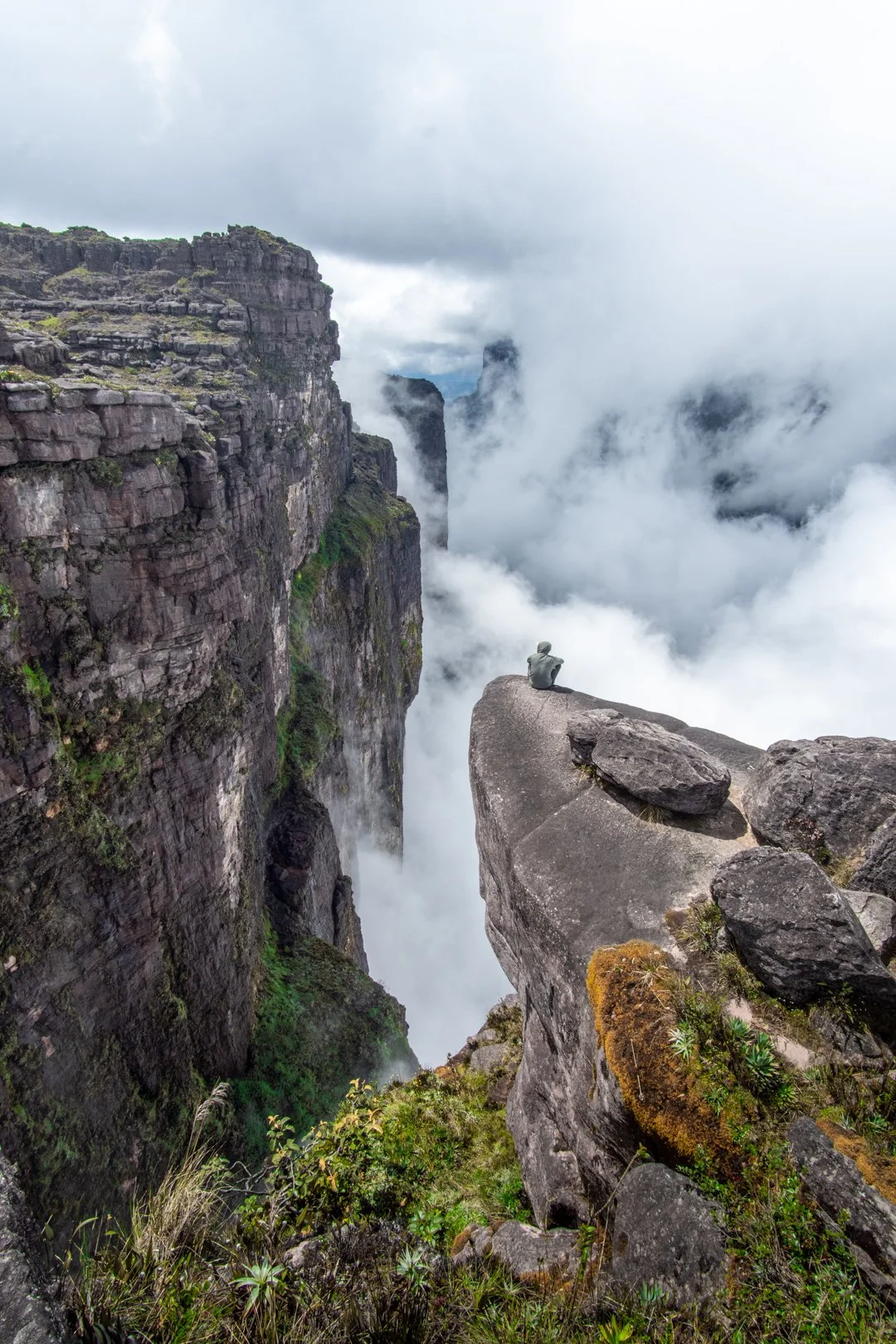 Person sitting on a large rock formation overlooking a deep canyon with cliffs and clouds on Tepui Roraima, Venezuela